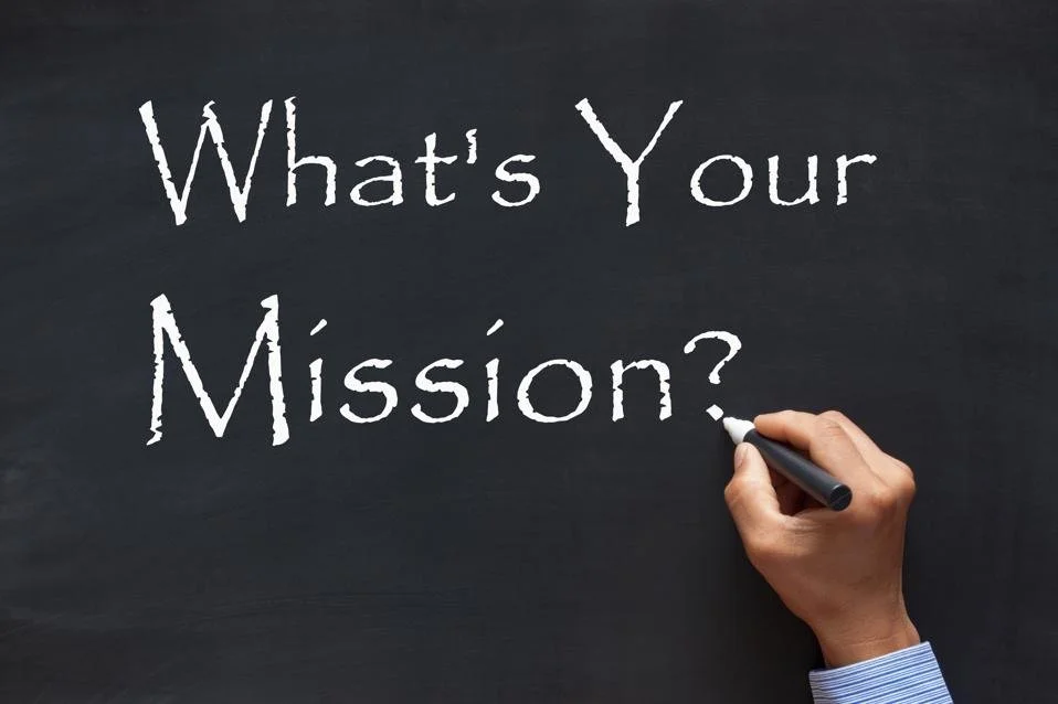 A person's hand writing on a blackboard with a white chalk, asking 'What's Your Mission?'