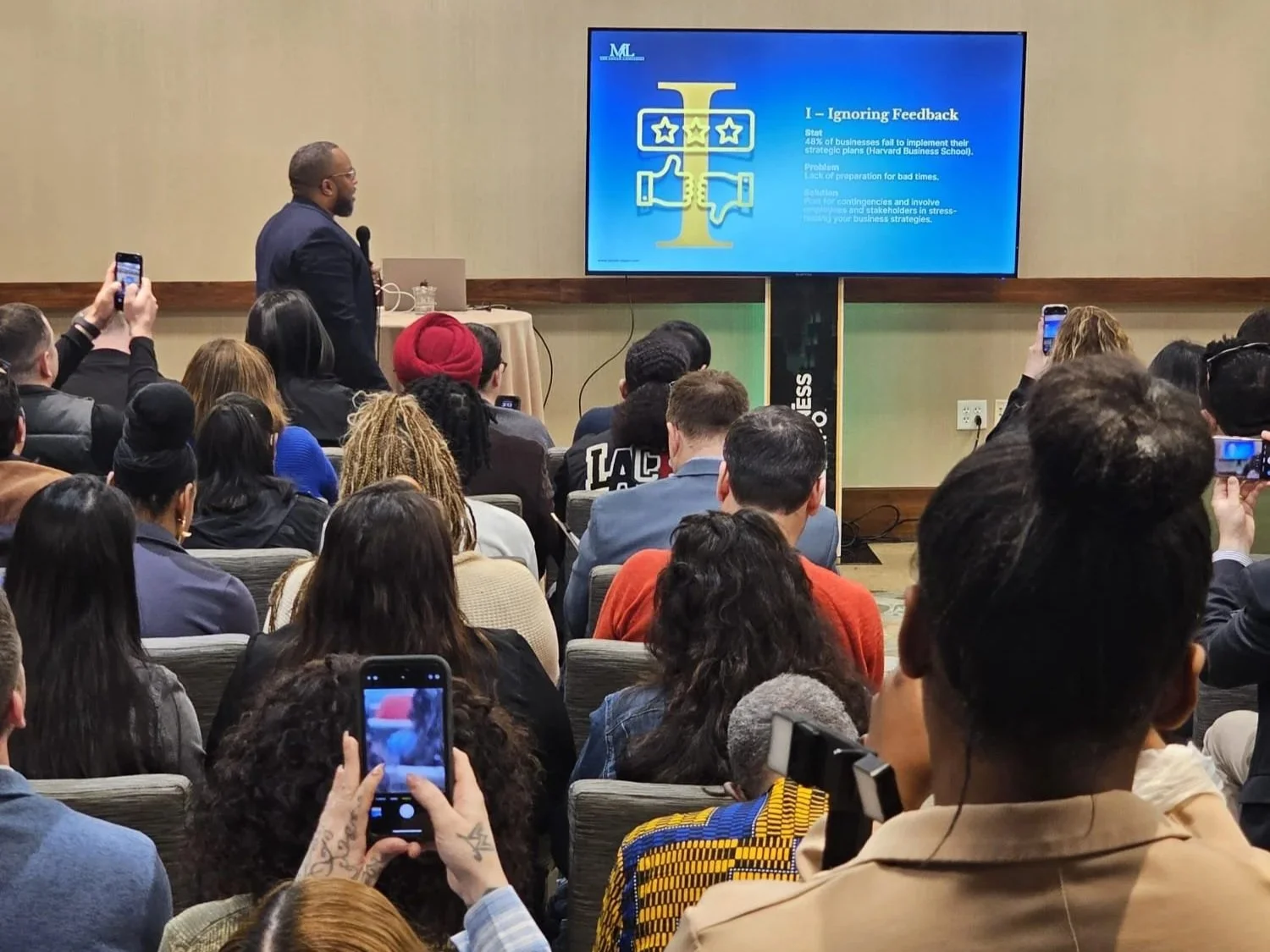 A man giving a presentation at a conference, standing near a large screen displaying a slide with the title "Ignoring Feedback." The slide includes text and a graphic of a balance scale with stars and a trophy. The audience is seated, many taking pictures or videos with smartphones.