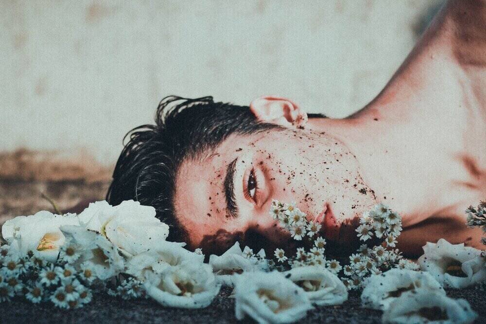 a white man lying on his side in the dirt with white flowers scattered on the ground and dirt on his face.