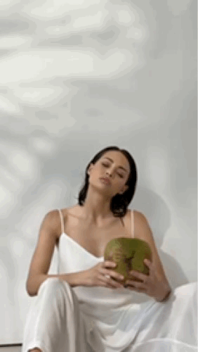 Petite women sitting under swaying palm shadow wearing sustainable petite tunic and trouser set, holding a jelly nut coconut under the sun.