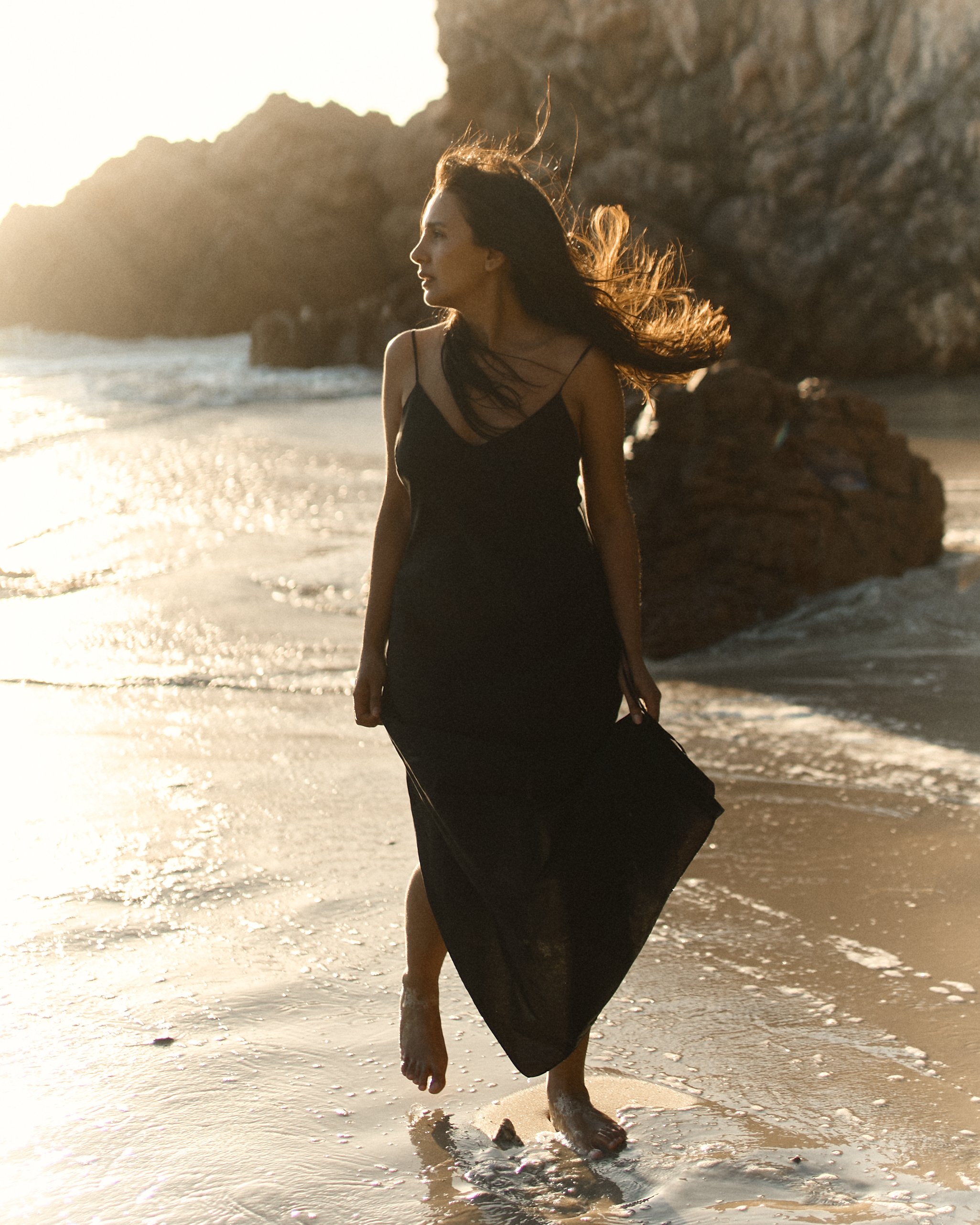 A woman with long hair in a black dress walking along the shoreline at sunset, with rocky cliffs in the background and waves gently crashing at her feet.