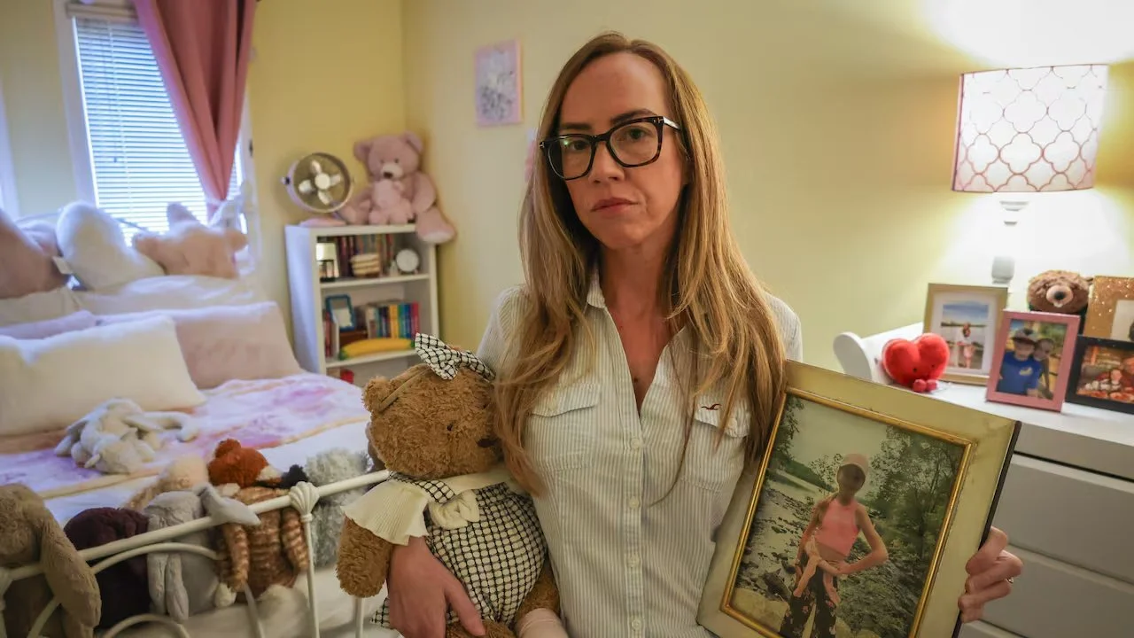 a woman looking serious, in a child's bedroom, holding a photo and a teddy bear.