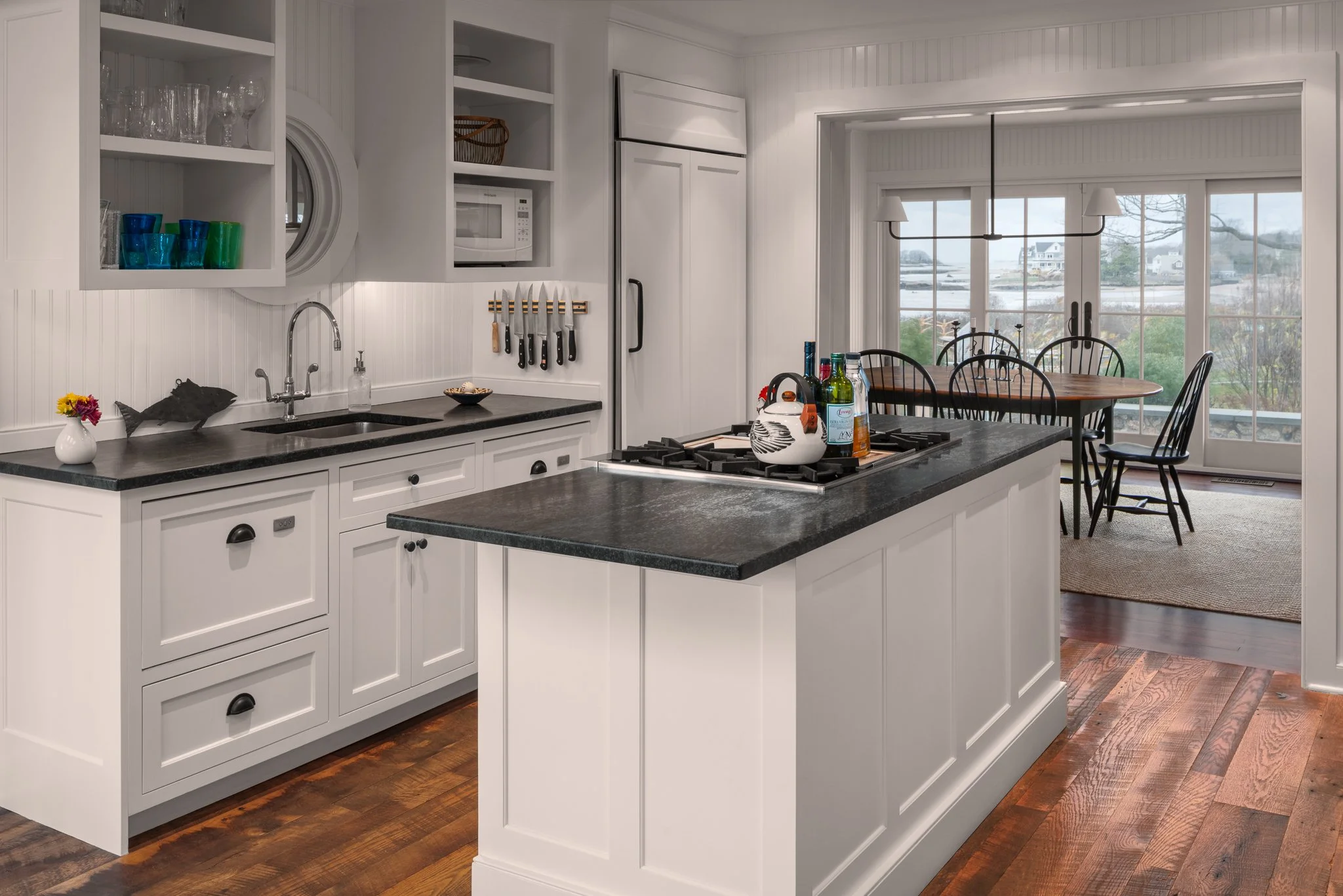 White kitchen with a cooktop island and granite countertops, the dining area and shoreline beyond.