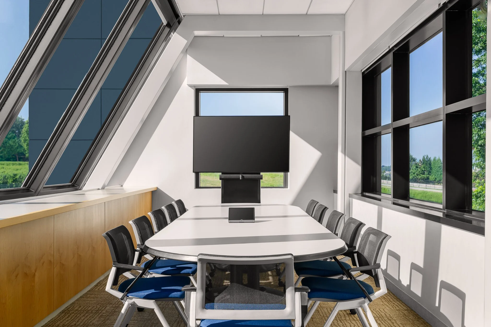Conference room under a slanted glass wall, with an oval table, blue-seated mesh chairs and a large display set in front of a window.
