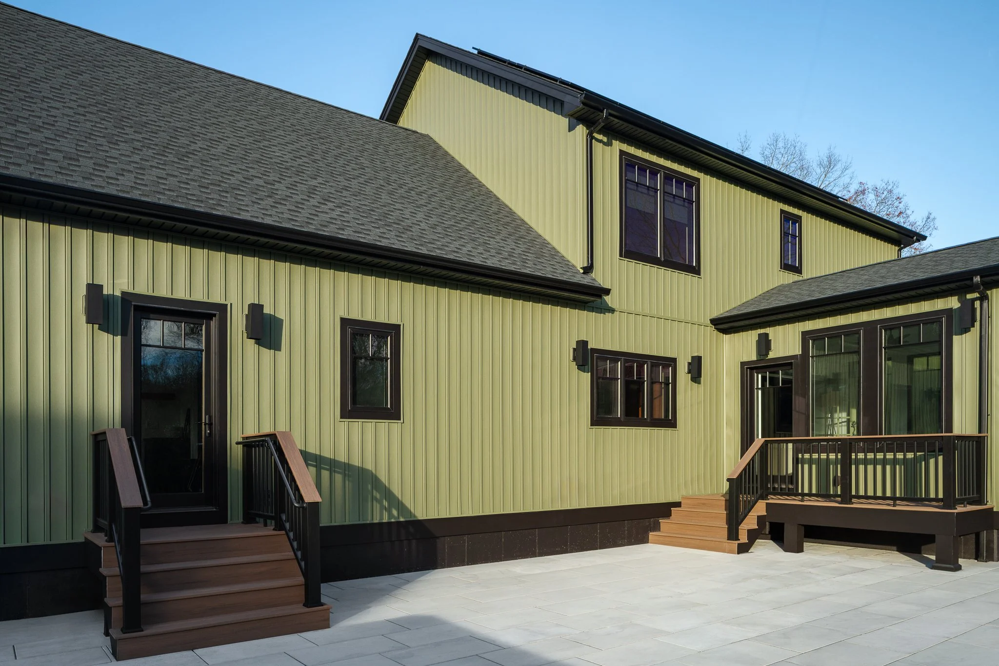 Rear exterior of the Westerly River Run home with sage green vertical siding, black-framed windows and composite steps leading down to a stone paver patio.