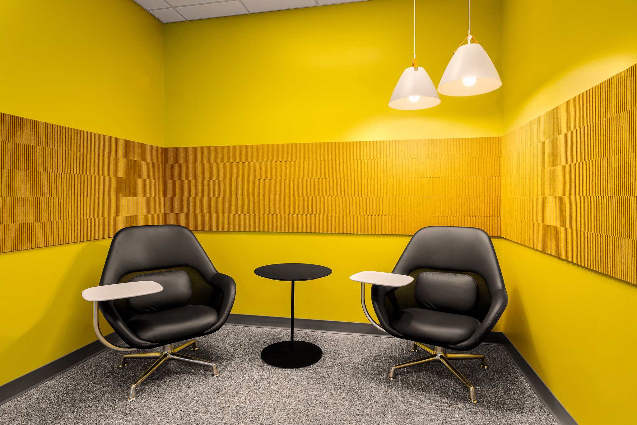 Yellow lounge nook with two black leather tablet-arm chairs, a small round table and acoustic wall panels.