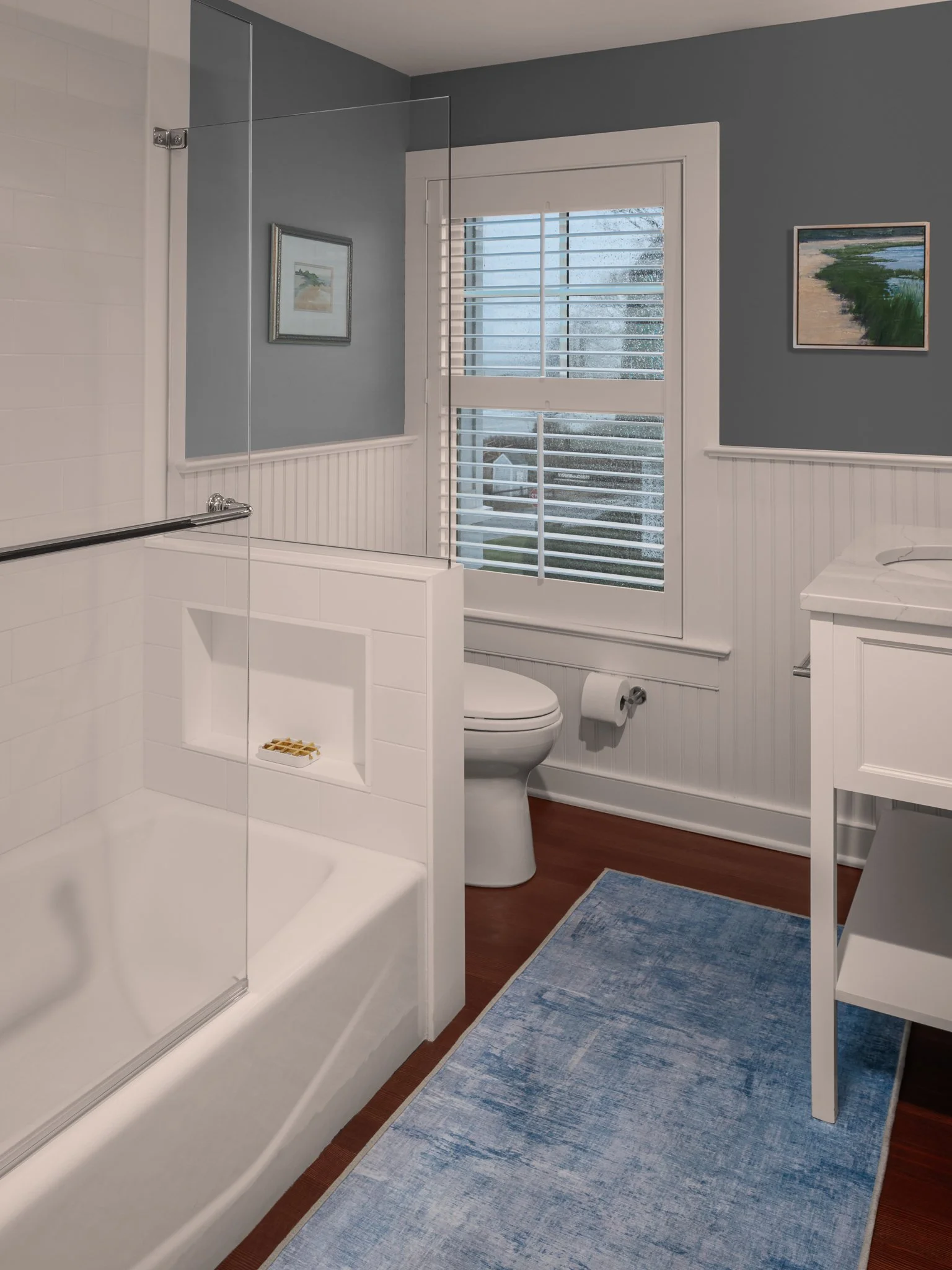 Compact bathroom with a tub, blue rug and rain-streaked window.