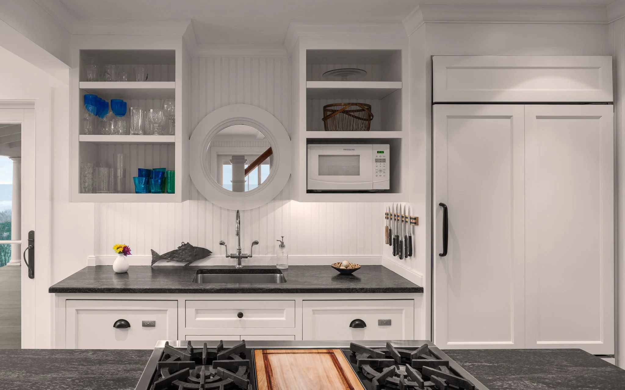 Kitchen island stovetop front and center, granite countertops, white walls, sink, cabinets with glassware and a circle window looking outside.