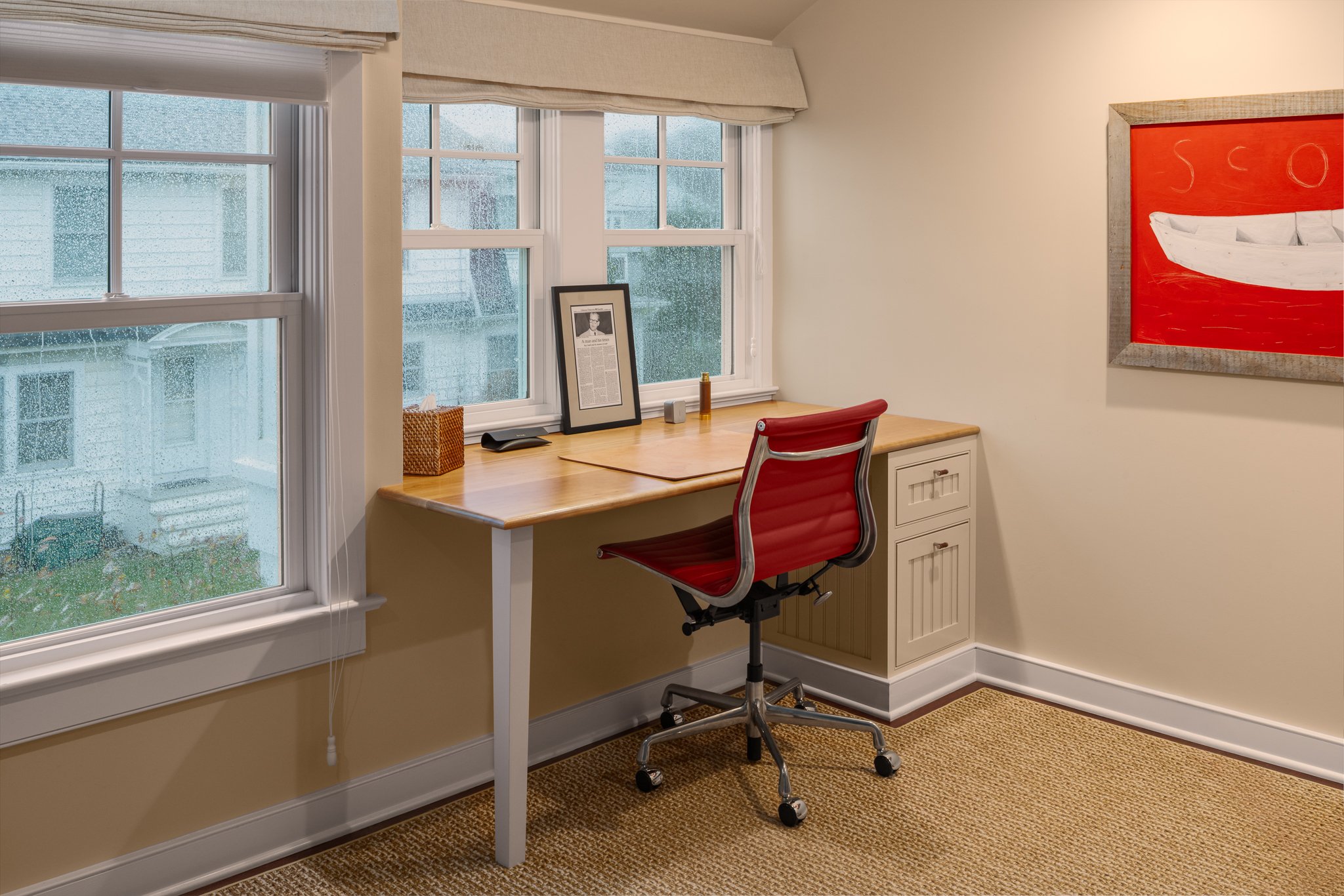 Writing desk under a window with a red leather chair and red artwork.