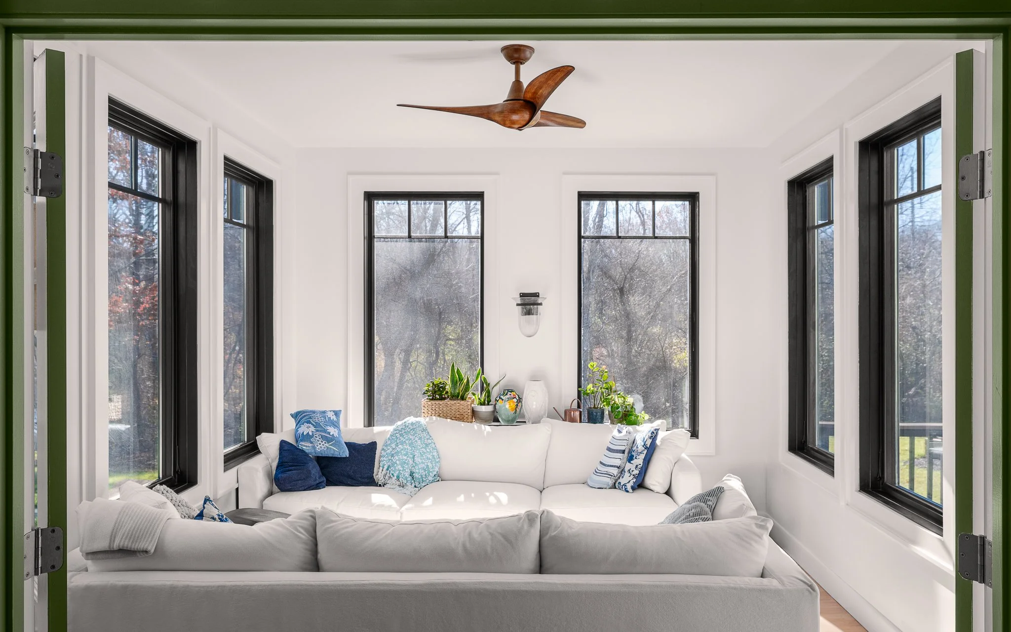 Sunroom wrapped in black-framed windows with white sofas and a wood-bladed ceiling fan.