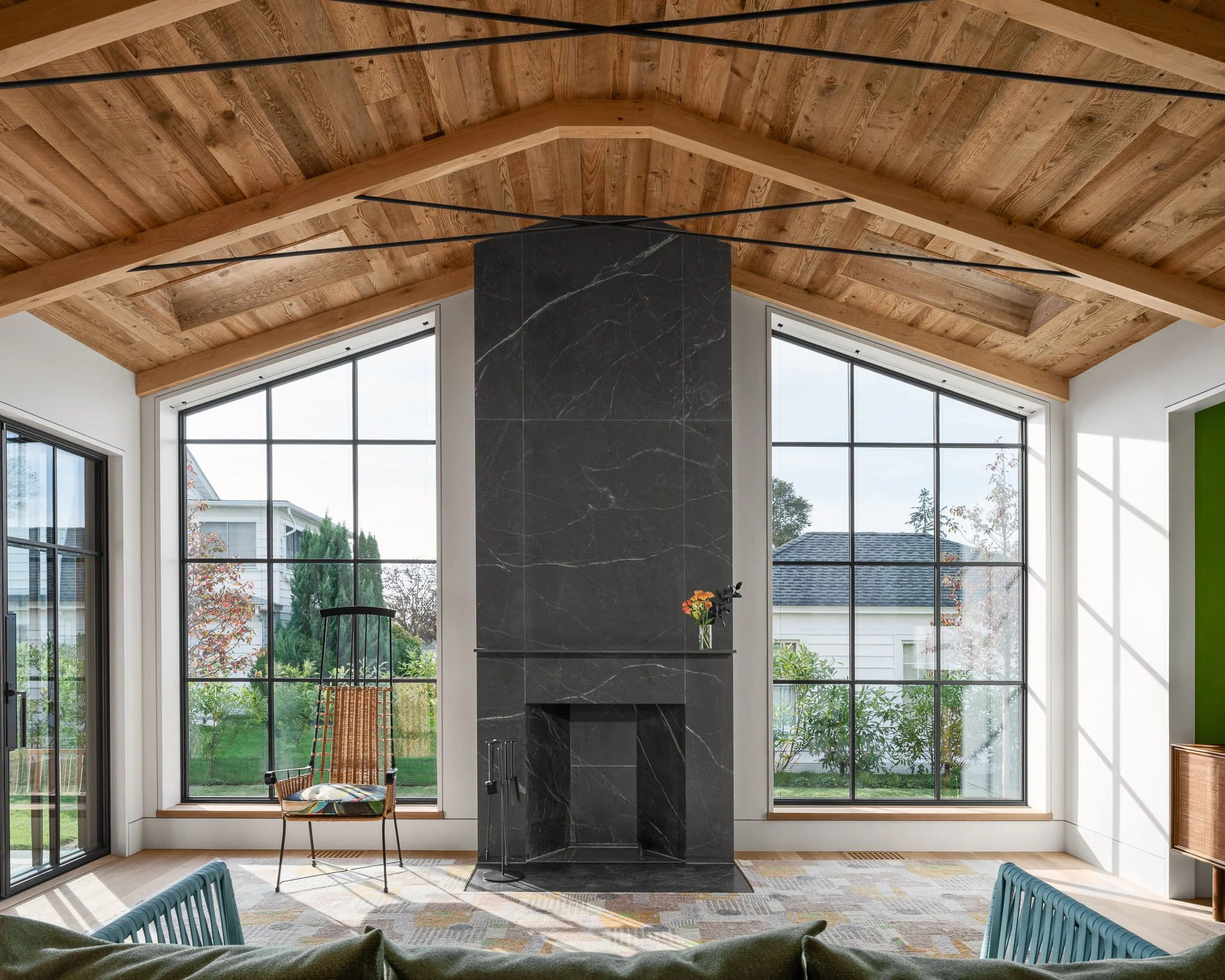 Great room with a dark stone fireplace rising into a wood ceiling between tall black-framed windows.