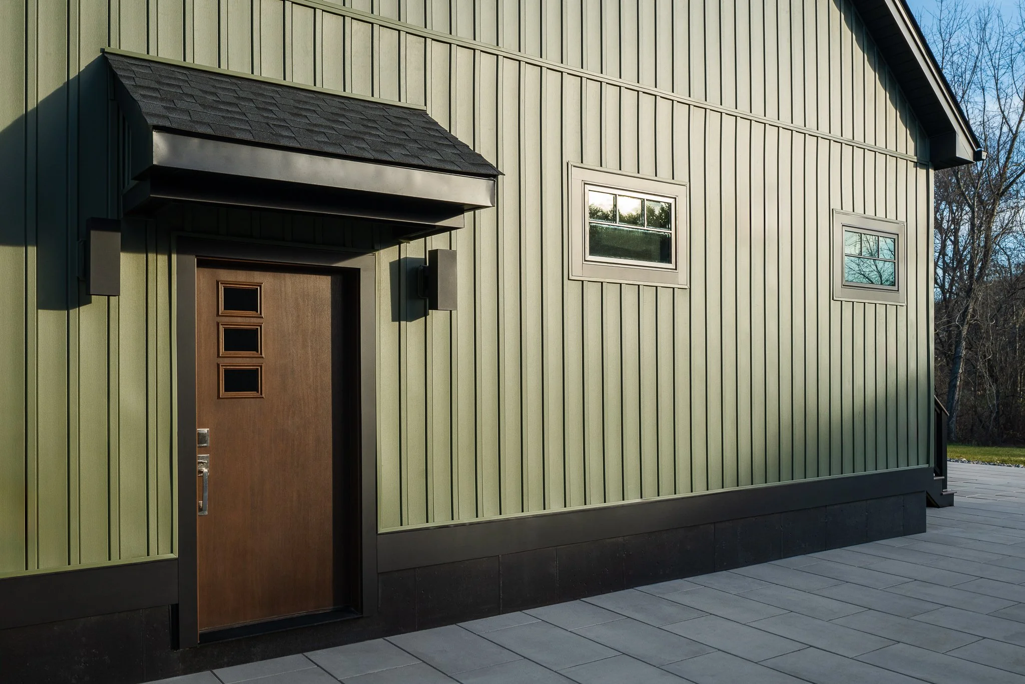 Wood-grain entry door with three small windows under a shingled overhang, set in lakeshore fern board-and-batten siding.