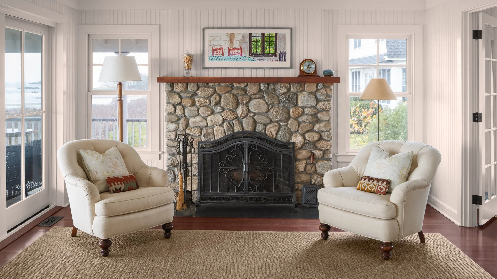 Fieldstone fireplace flanked by two white armchairs and water-view windows.