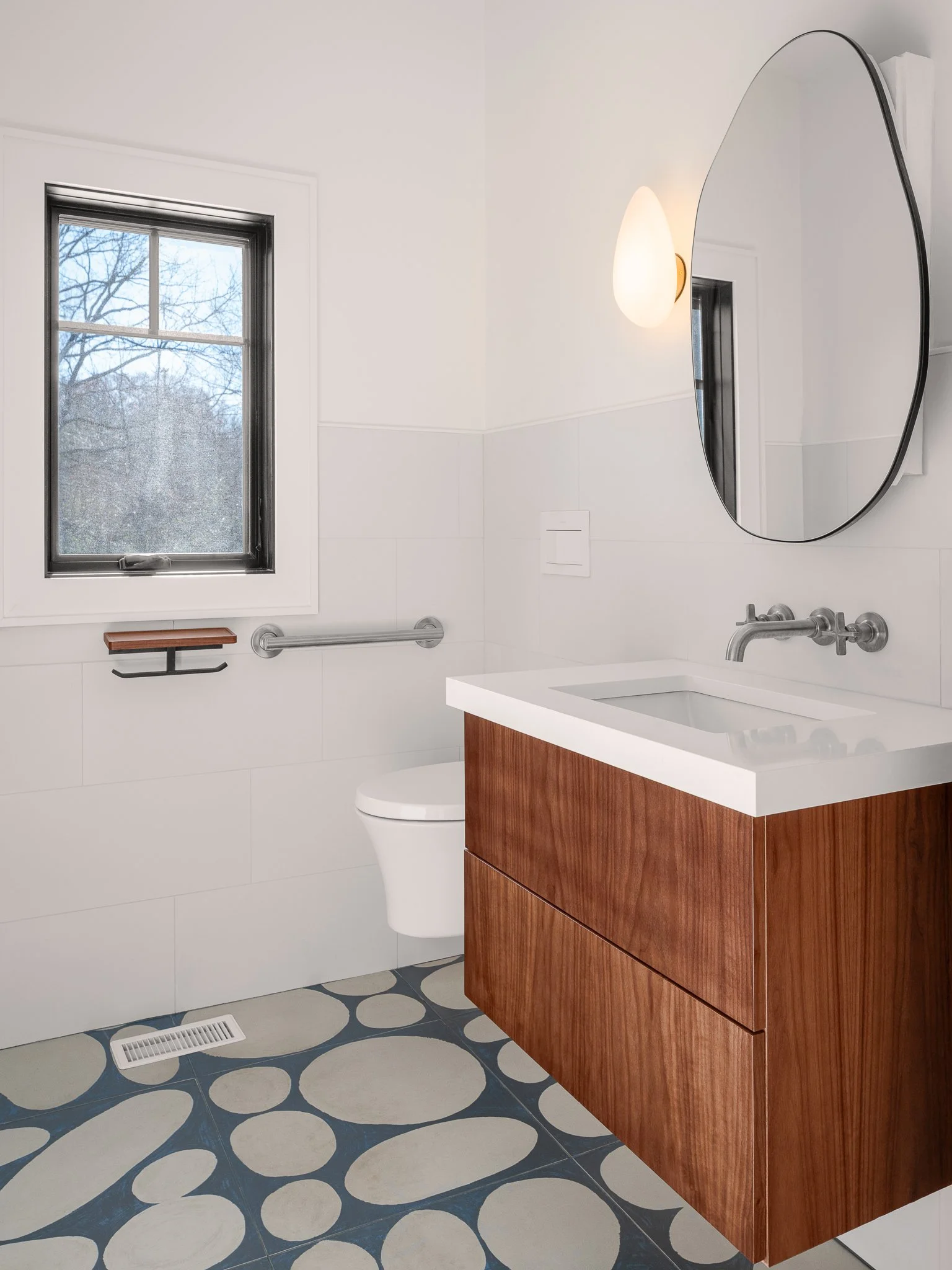 Powder room with a pebble tile floor, floating wood vanity and oval mirror.