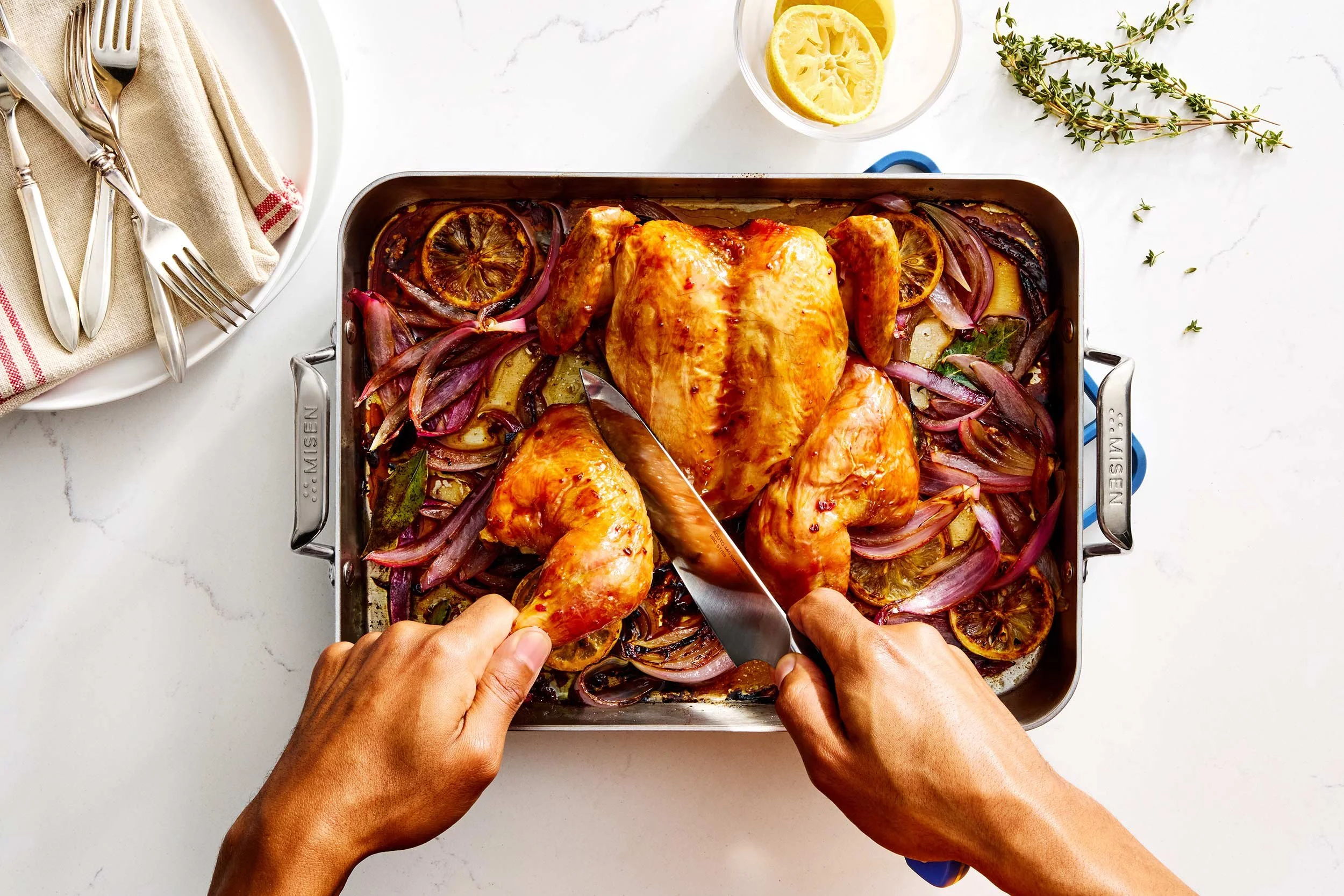 Baked chicken with roasted lemons and vegetables in a metal baking dish, with hands carving into the chicken, set on a white surface with a lemon glass and a utensil plate nearby.