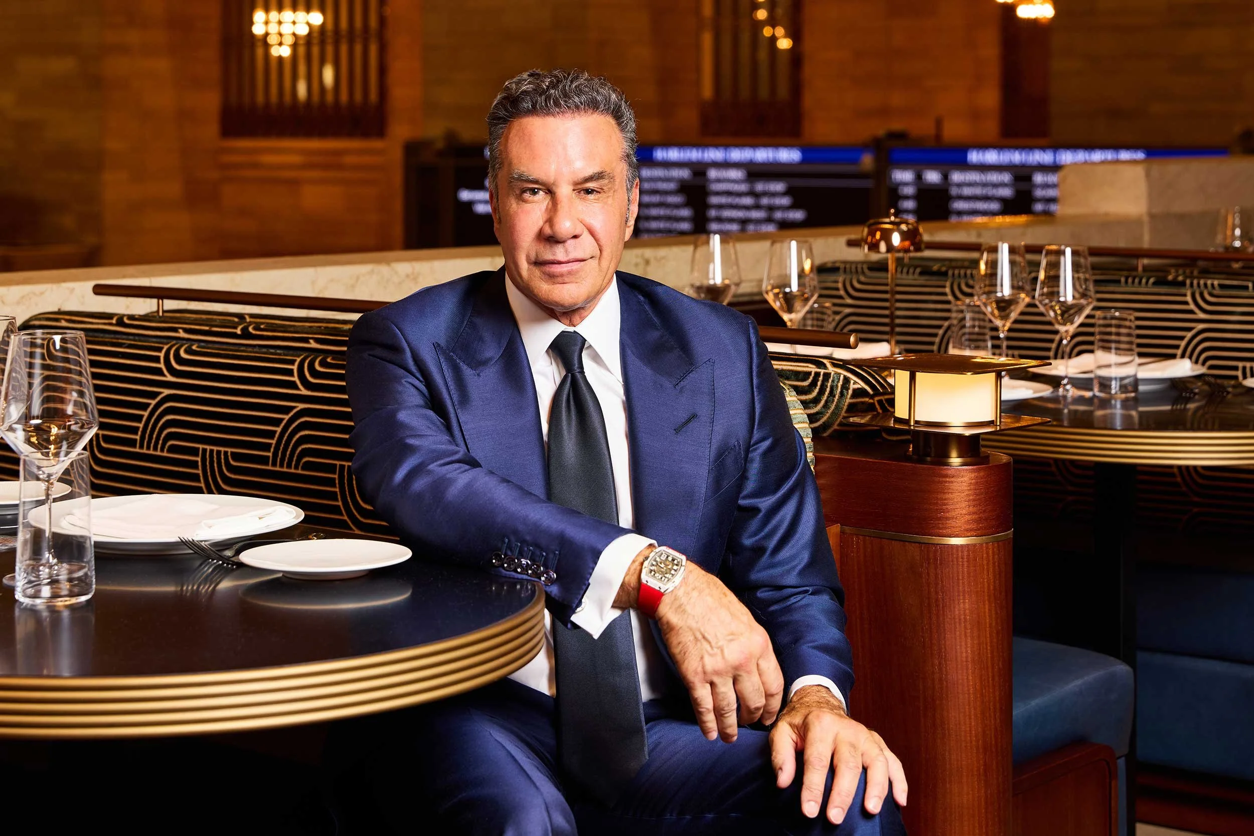 Joseph Palladino in a blue suit sits at a restaurant table with a serious expression. The table is set with plates, glasses, and utensils, with a bar or display in the background.