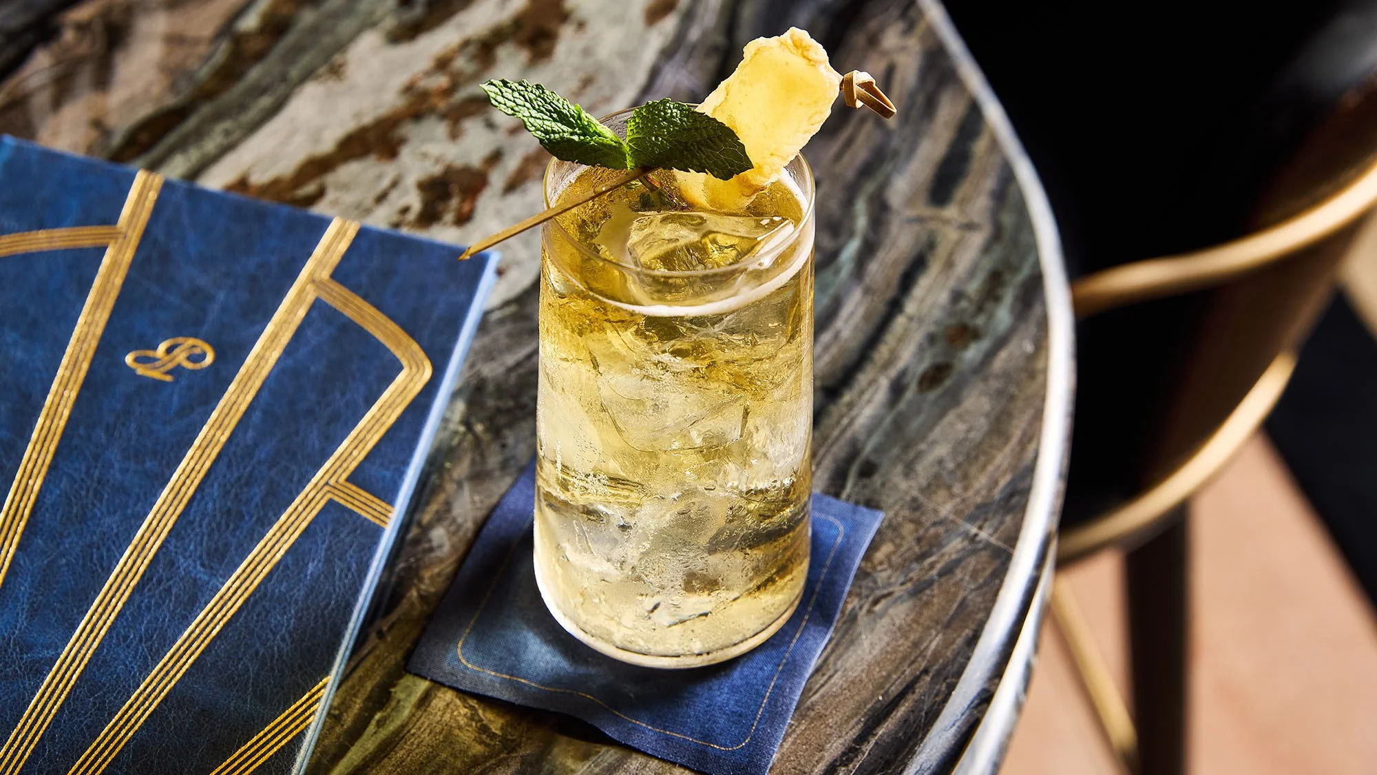 A tall glass of a yellowish cocktail with ice, garnished with a pineapple slice and a fresh mint sprig, on a wooden table.