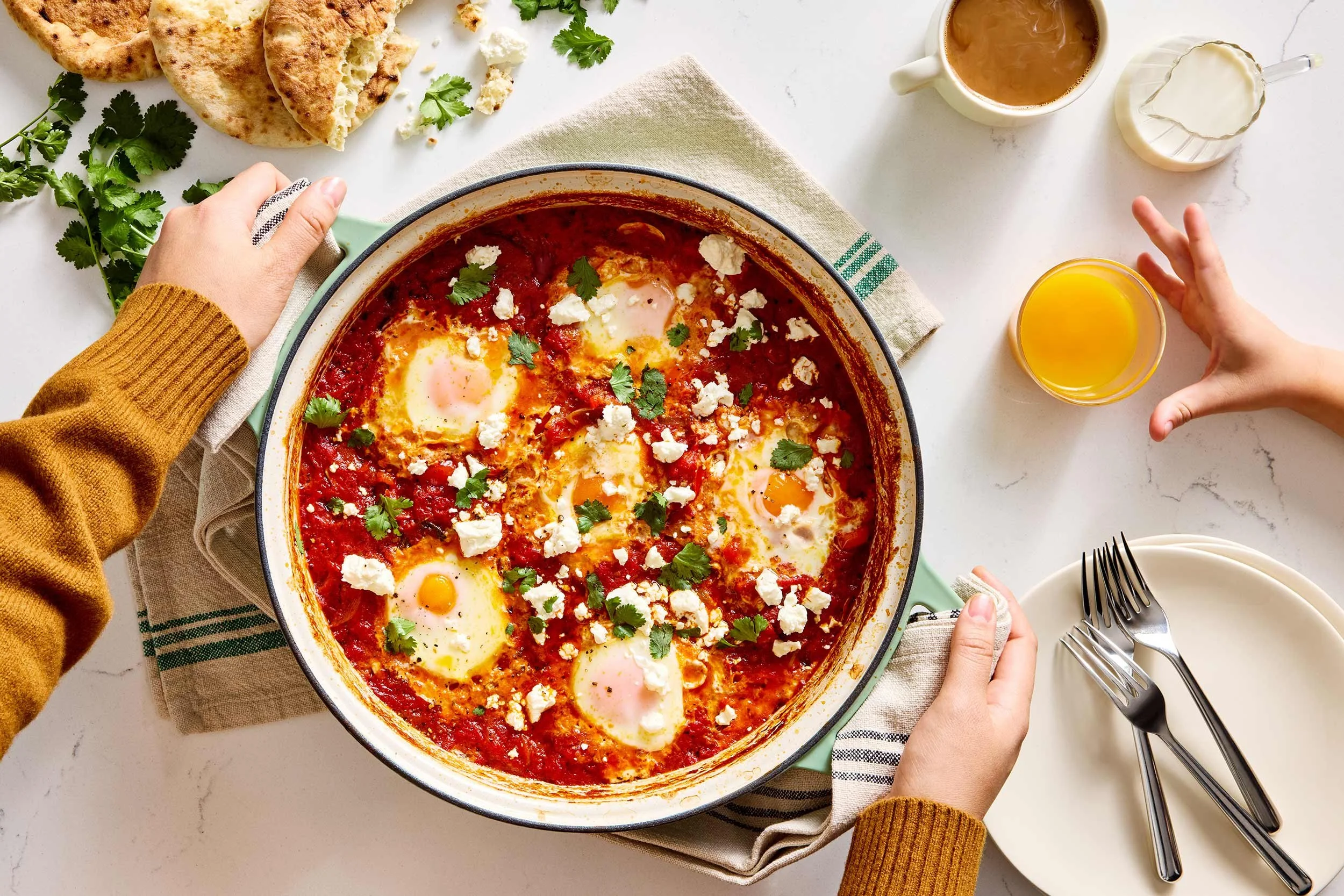 A person holding a large Misen skillet of shakshuka, topped with crumbled feta cheese and cilantro. There are slices of bread, a cup of coffee, a glass of orange juice and a child's hand, and utensils on plates around the skillet.
