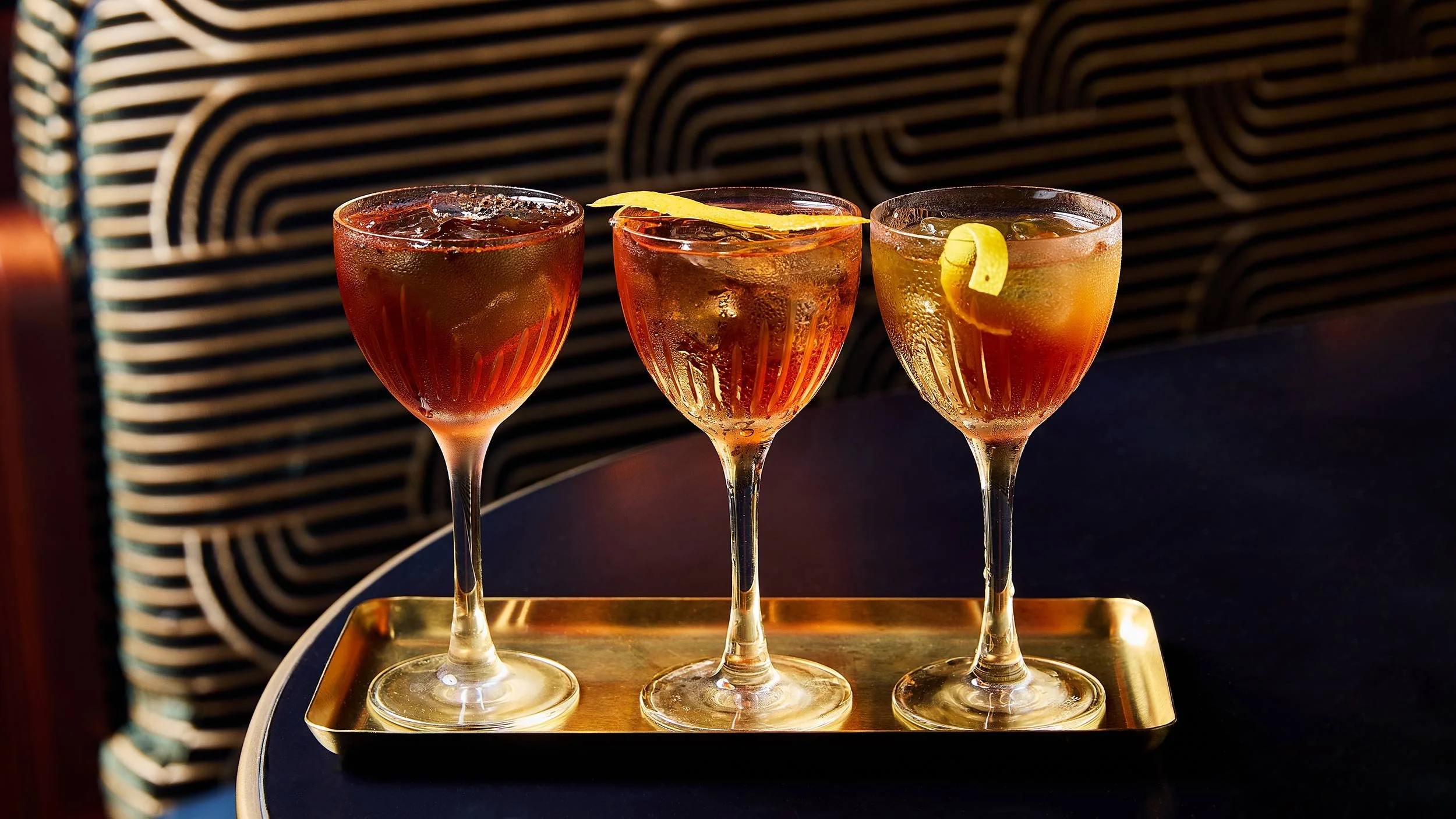 Three cocktails in coupe glasses on a gold tray, each garnished with lemon peels, set on a dark table against a patterned background.