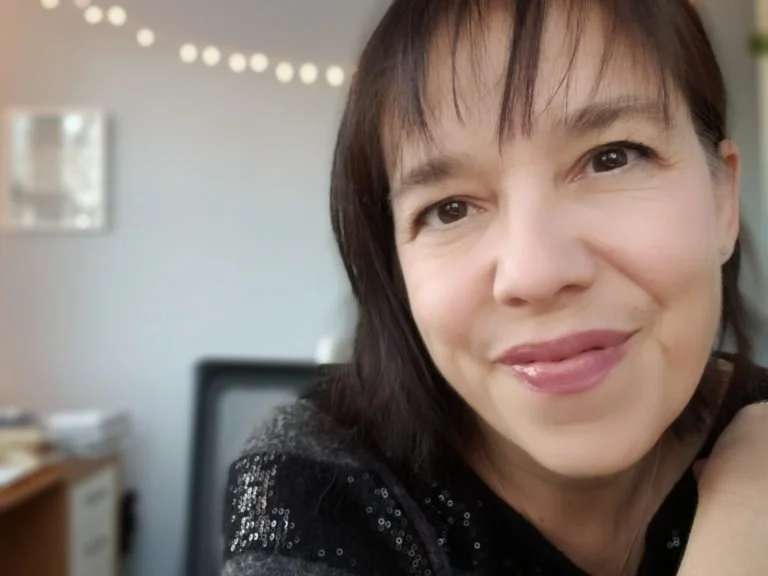 Creative coach, artist and writer Jamie Ridler smiling in her studio, wearing a dark sweater, with blurred background featuring sparkling lights.