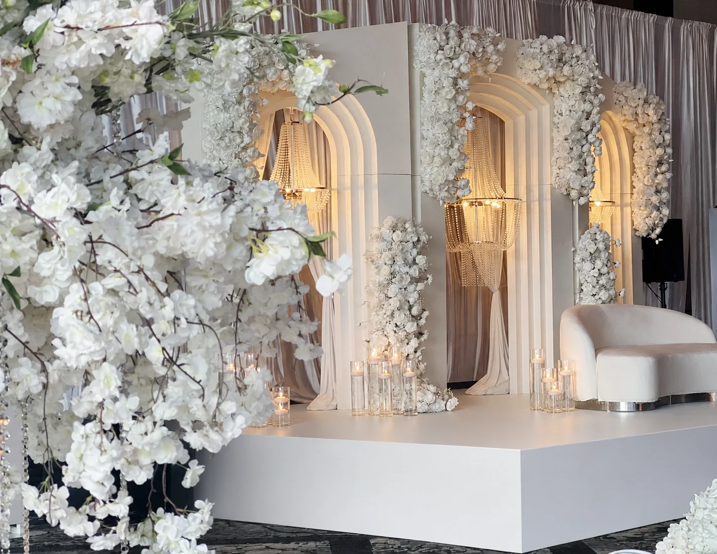 A luxurious wedding stage featuring tall white pillar-style arches decorated with cascading white flowers, elegant crystal chandeliers, and a sleek white loveseat, creating a modern and sophisticated ambiance.
