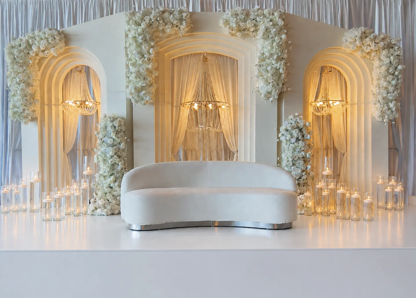 A stunning wedding stage with tall white arch structures adorned with white florals and crystal chandeliers, complemented by a modern white sofa — creating a bright, elegant, and timeless setup.
