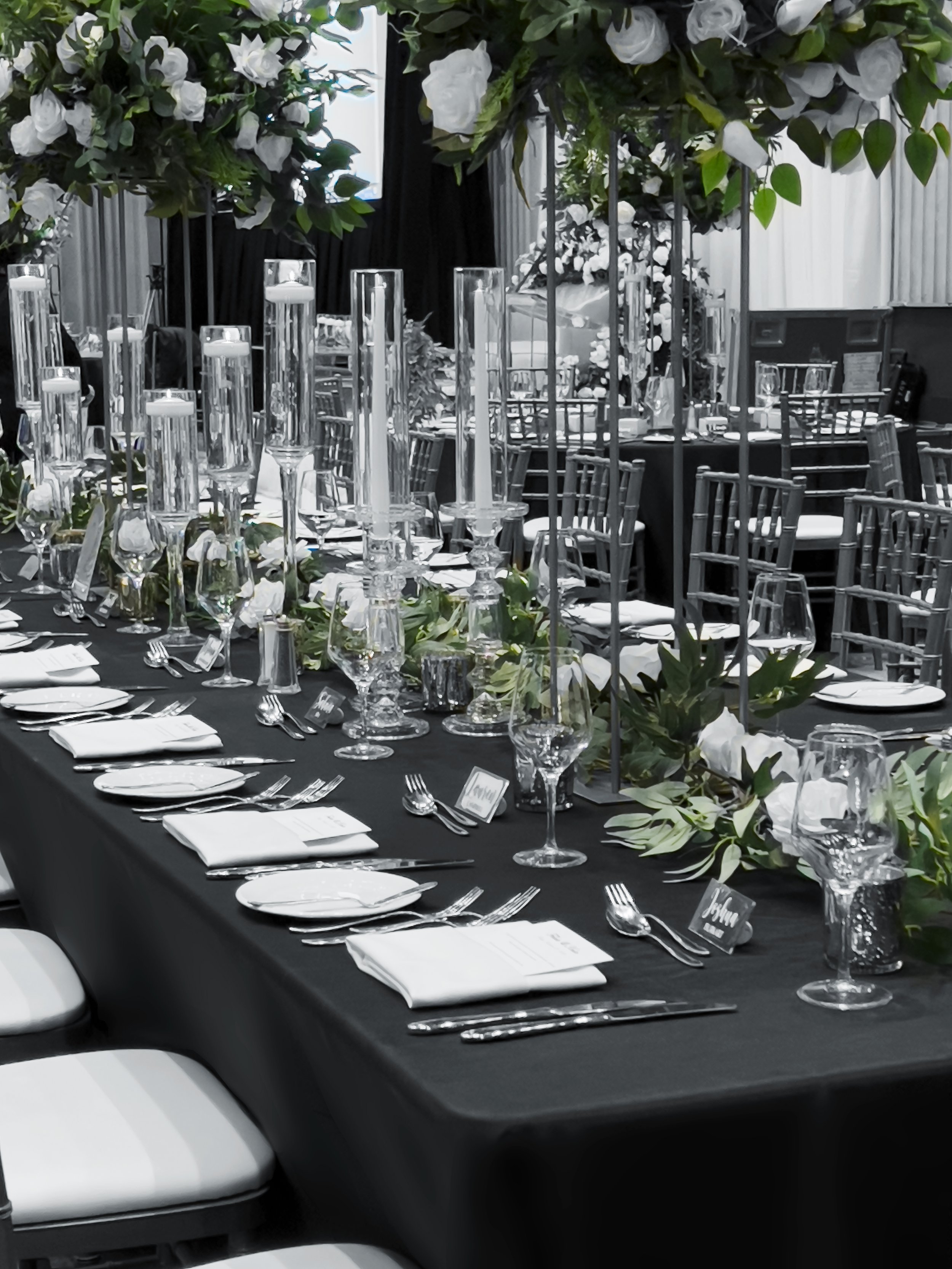 Elegant indoor event table setup with a black tablecloth, white napkins, glassware, silverware, floral arrangements, and candles.