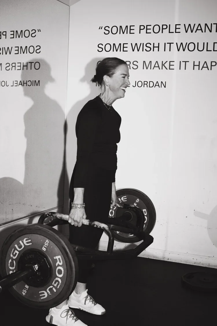 Sarah Schmidt in athletic wear smiles while holding a barbell loaded with Rogue weights. Motivational quotes are on the wall. Energetic and empowering tone.