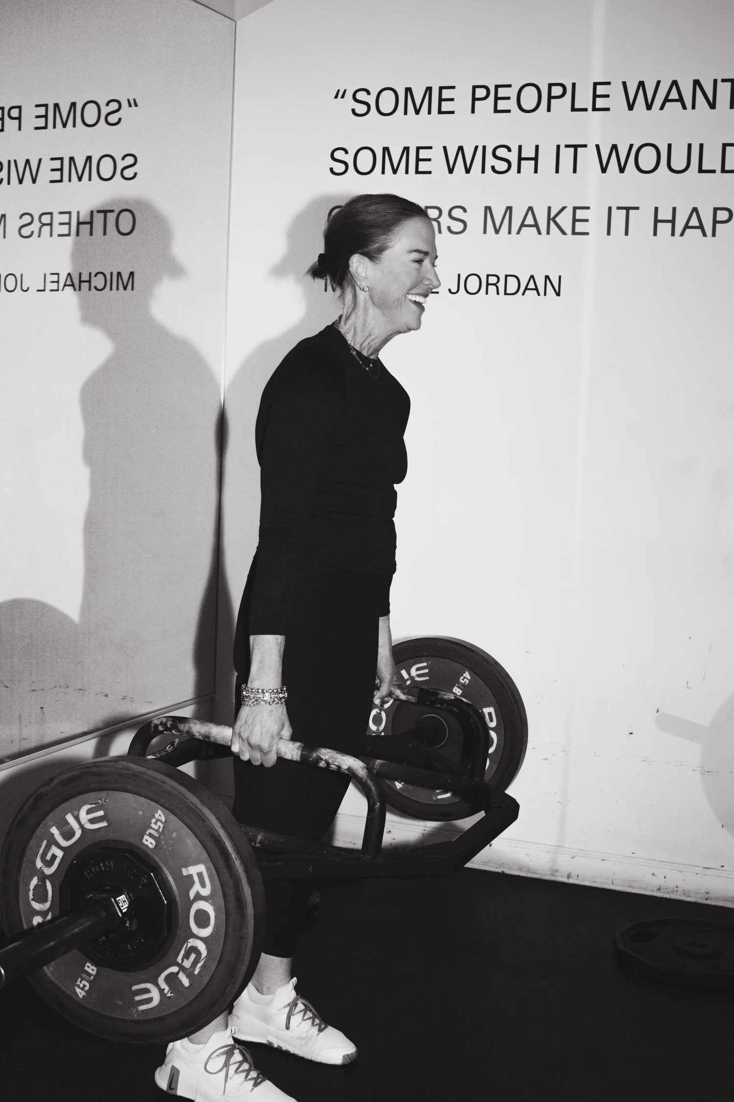 Sarah Schmidt in athletic wear smiles while holding a barbell loaded with Rogue weights. Motivational quotes are on the wall. Energetic and empowering tone.
