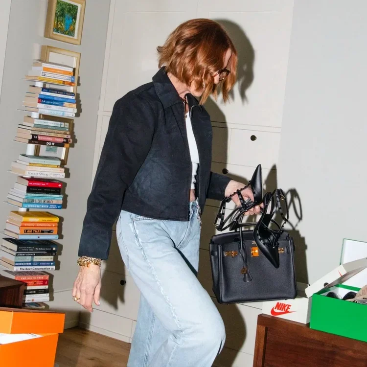 Sarah Schmidt wearing jeans and a jacket holds a bag, standing confidently with a neutral background.