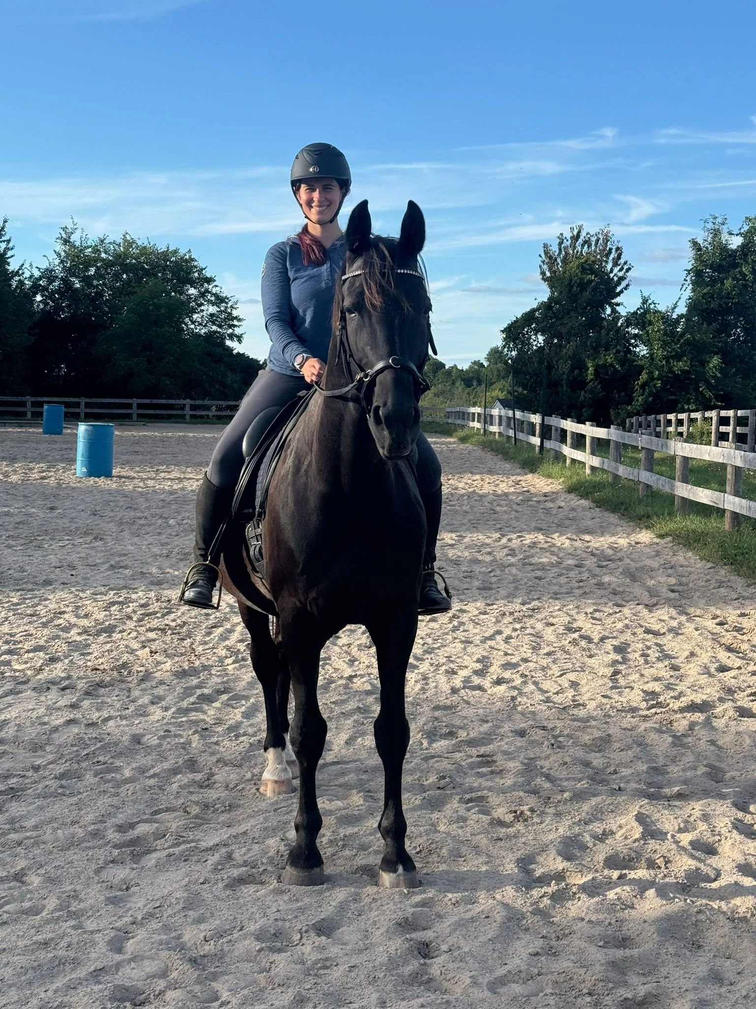 A woman riding a black horse in an outdoor riding arena, wearing a riding helmet and casual riding clothes, with trees and a blue sky in the background.
