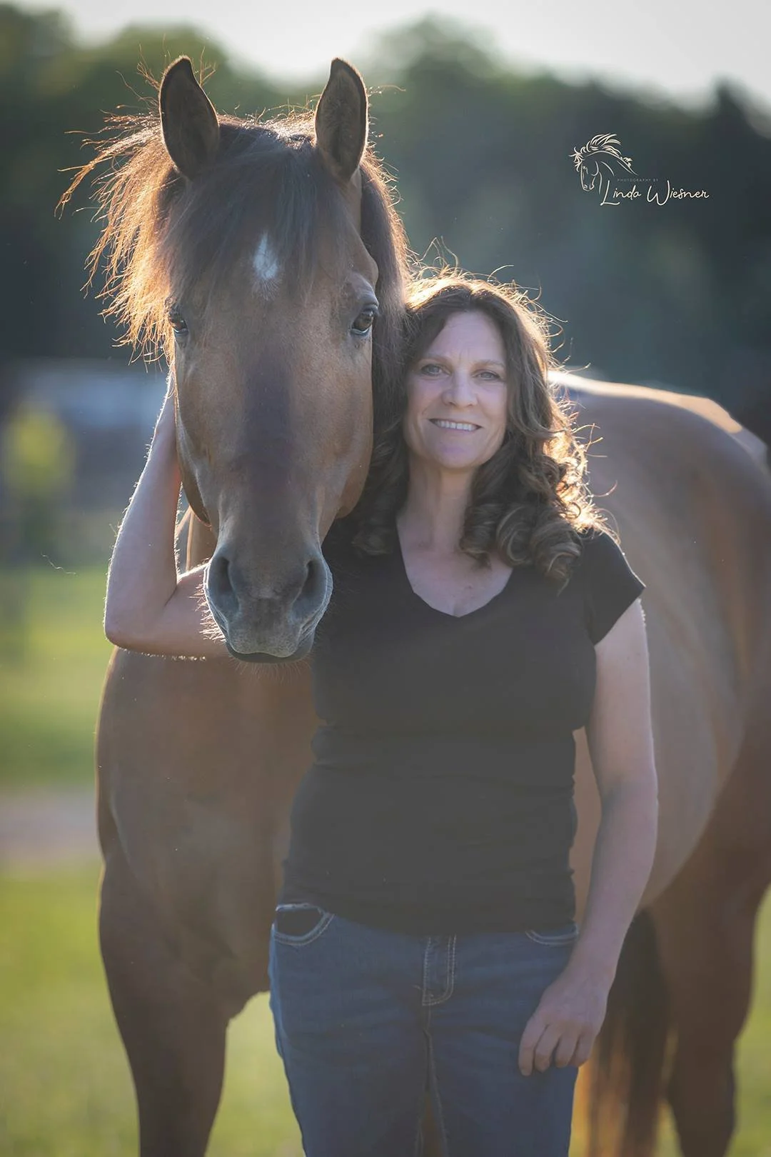 A woman standing outdoors next to a brown horse, smiling, with sunlight backlighting. The woman has curly brown hair, wearing a black t-shirt and blue jeans.