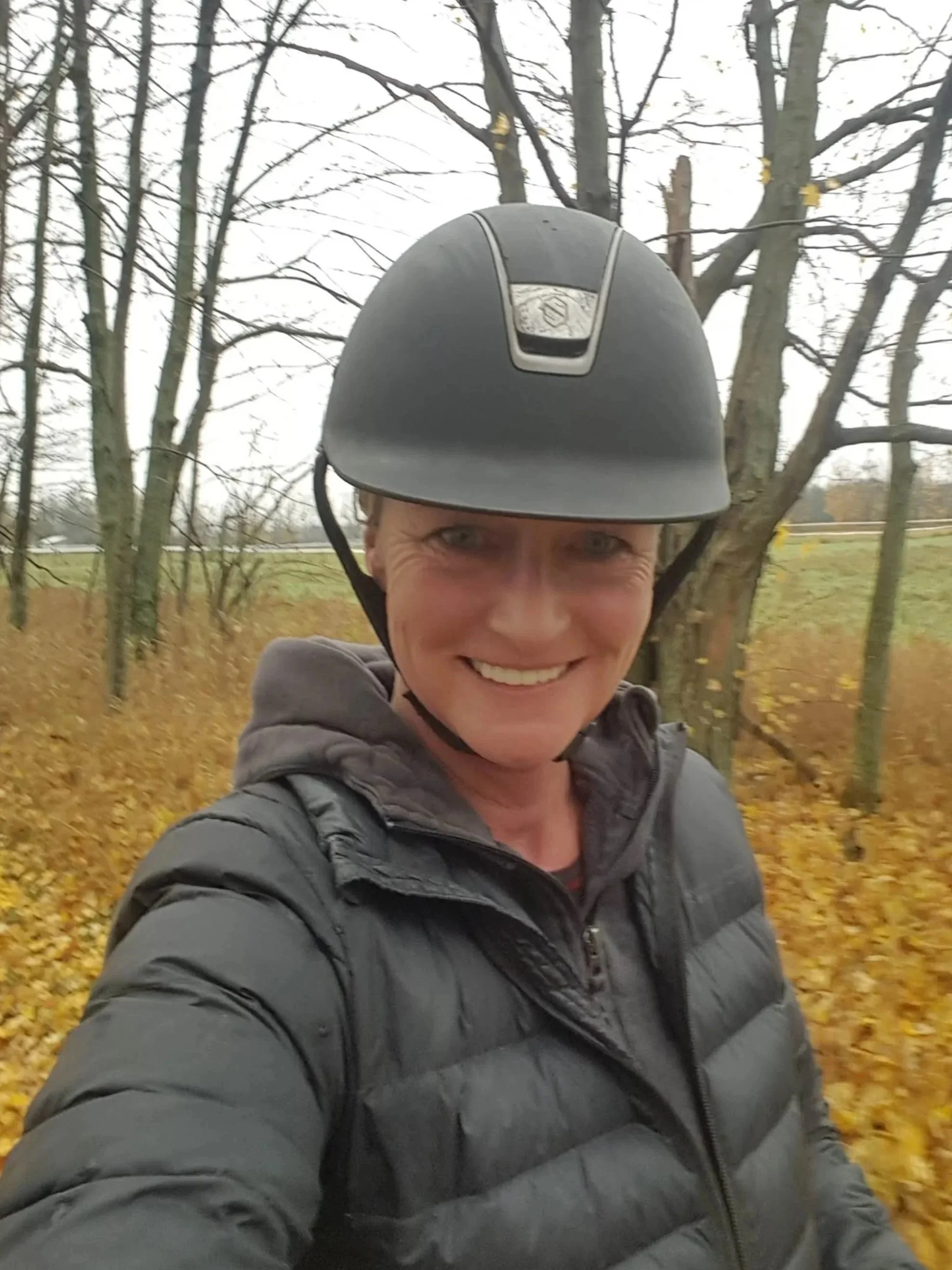 A rider and coach wearing a black helmet and a gray jacket taking a selfie outdoors in autumn, with trees and yellow fallen leaves in the background.