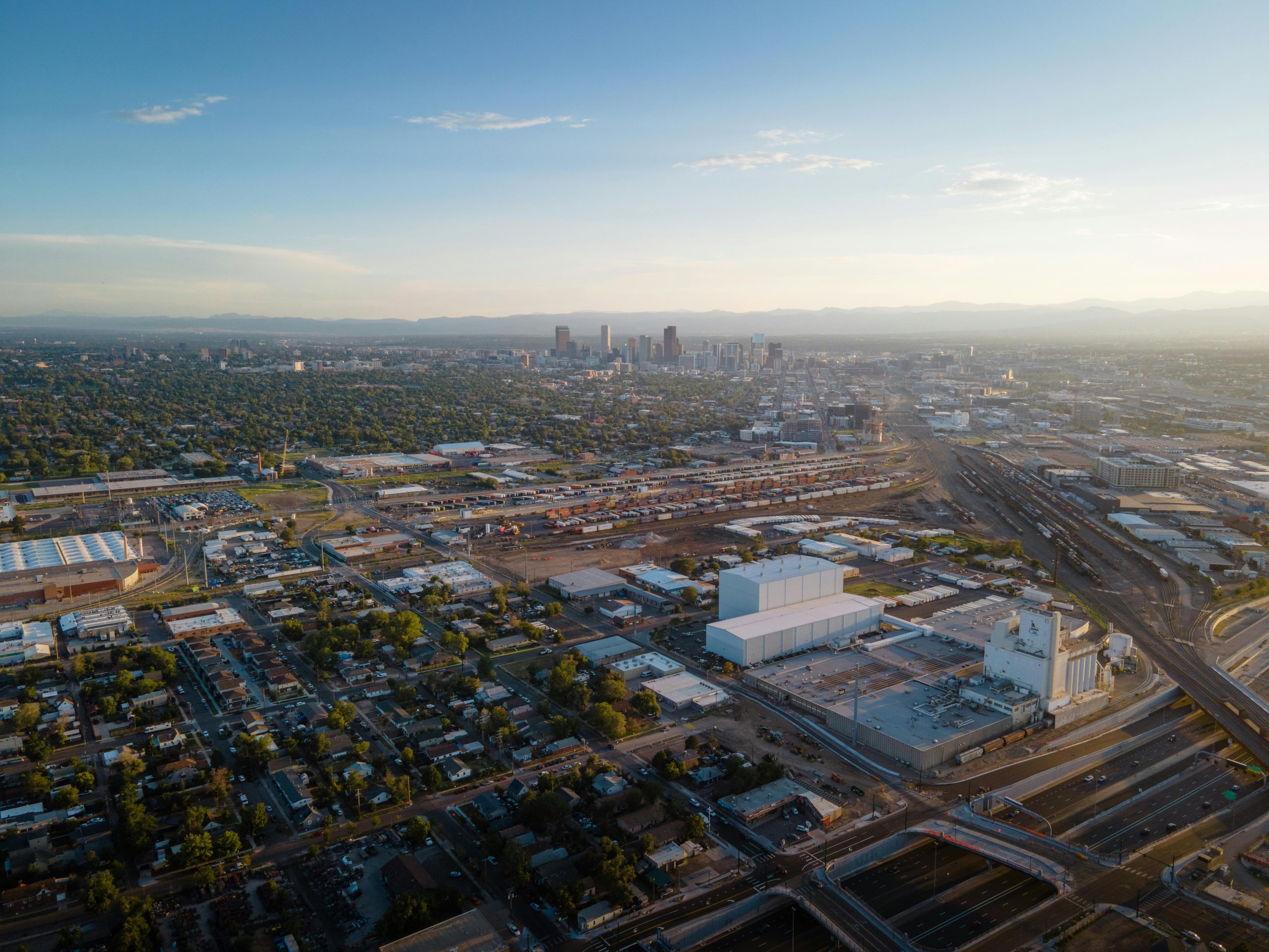 Aerial view of a cityscape during sunset showing residential neighborhoods, industrial areas, train tracks, and downtown skyscrapers in the distance.
