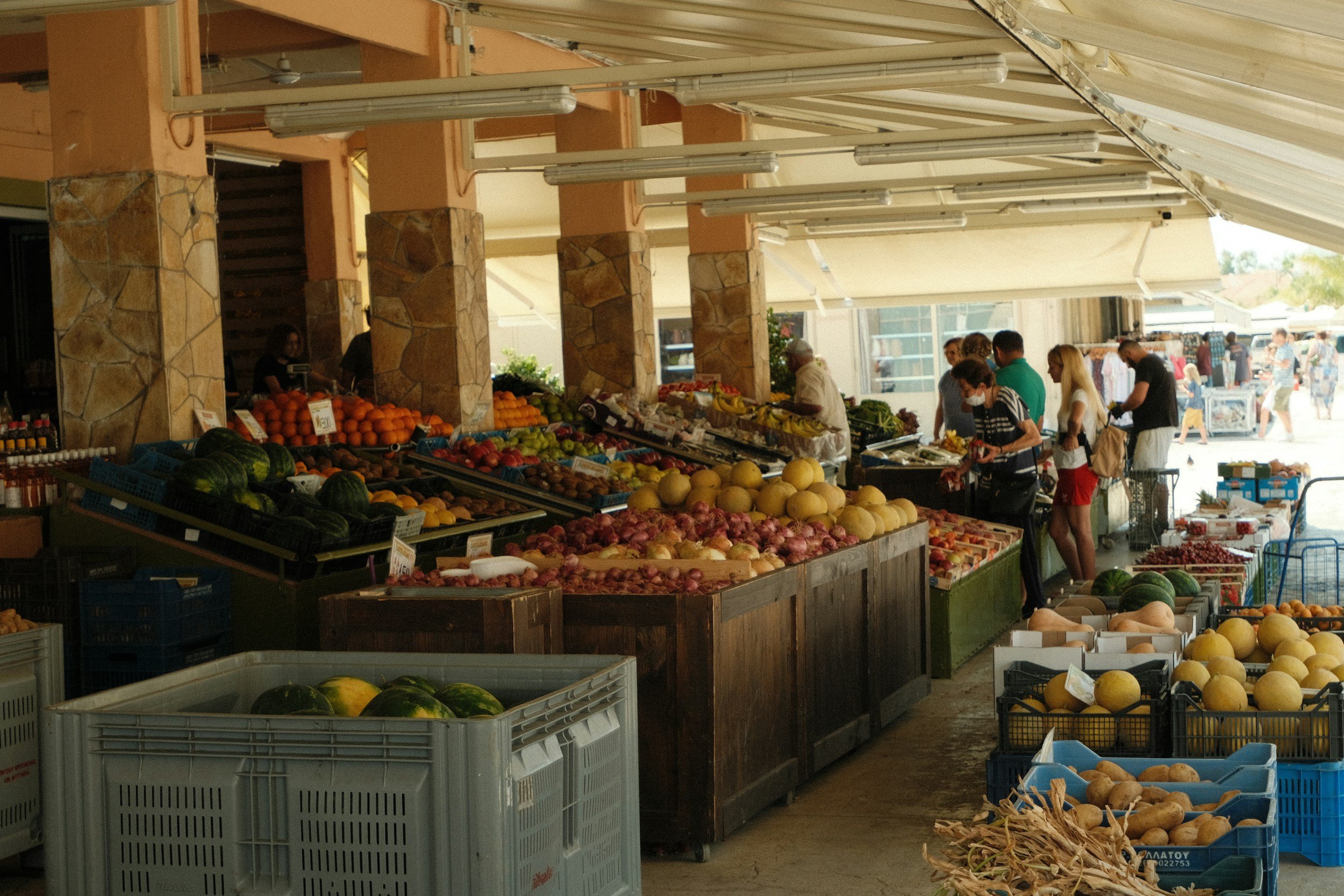 People shopping at an outdoor fruit market with various fresh fruits displayed, including watermelons, melons, and onions, under a beige canopy with stone pillars.