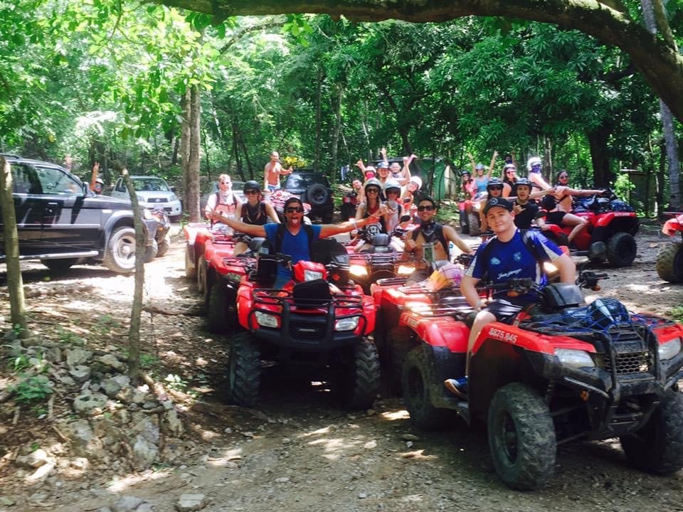 Group of people riding red ATVs in a forest trail with trees overhead.