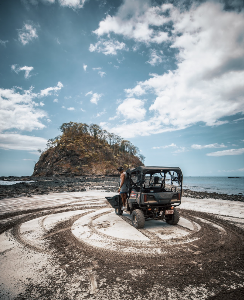 Man standing next to an off-road vehicle on a sandy beach, with circular tire tracks and a rocky island in the background. The sky is partly cloudy.