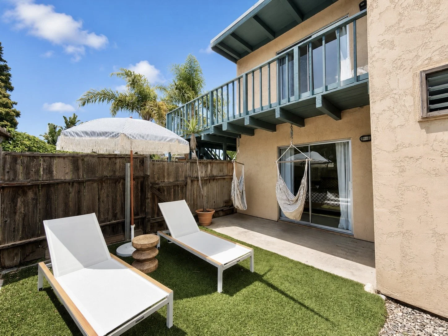 Outdoor patio with two wooden sun loungers, striped umbrella, two hanging hammocks, and potted palm tree on artificial grass and concrete. Adjacent wood fence and building with balcony.