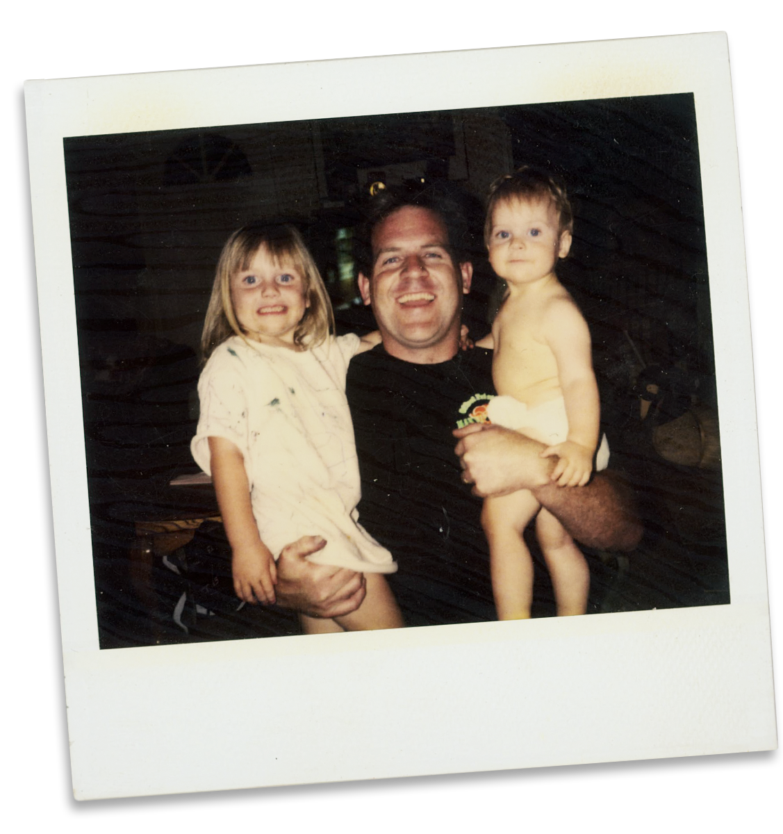 A man holding two young children, one girl and one boy, in a rustic indoor setting.