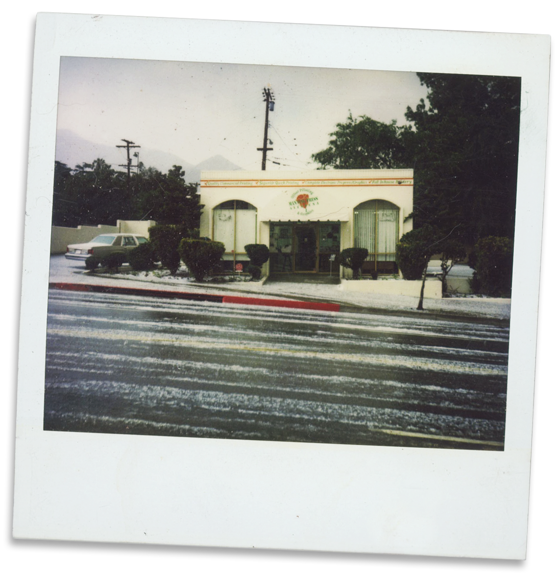 A vintage photo of a small restaurant with a white facade, three windows, and an entrance, surrounded by bushes, with a car parked on the street and utility poles in the background.