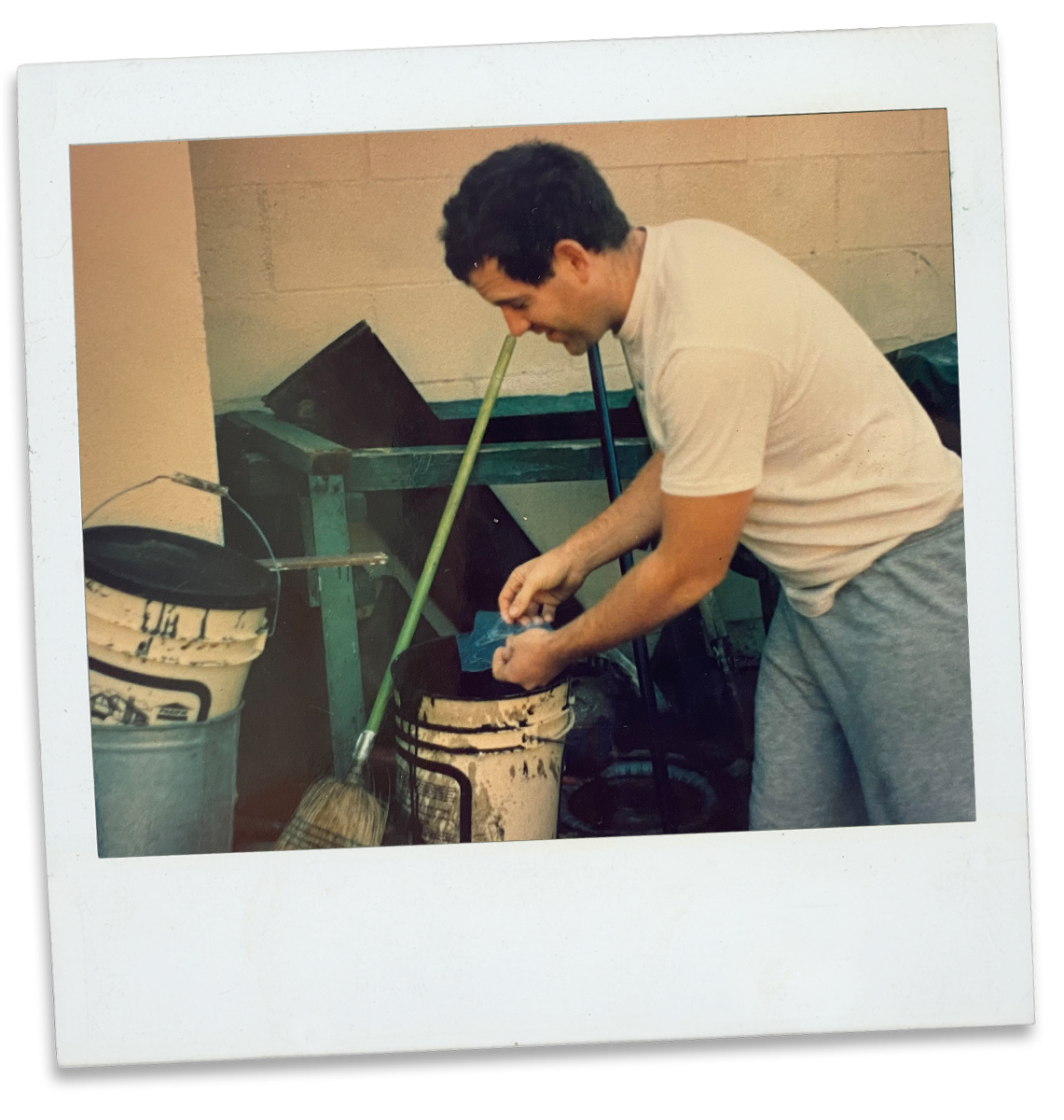 A man in a white t-shirt and gray sweatpants standing over a paint bucket, working on a project in a workshop with tools and paint buckets around.