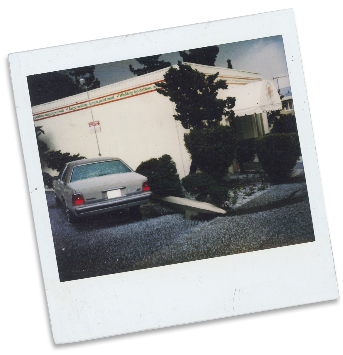 A Polaroid photo of a silver car parked in front of a white building with a large awning. There are bushes and trees around the building, and a street with gravel in the foreground.