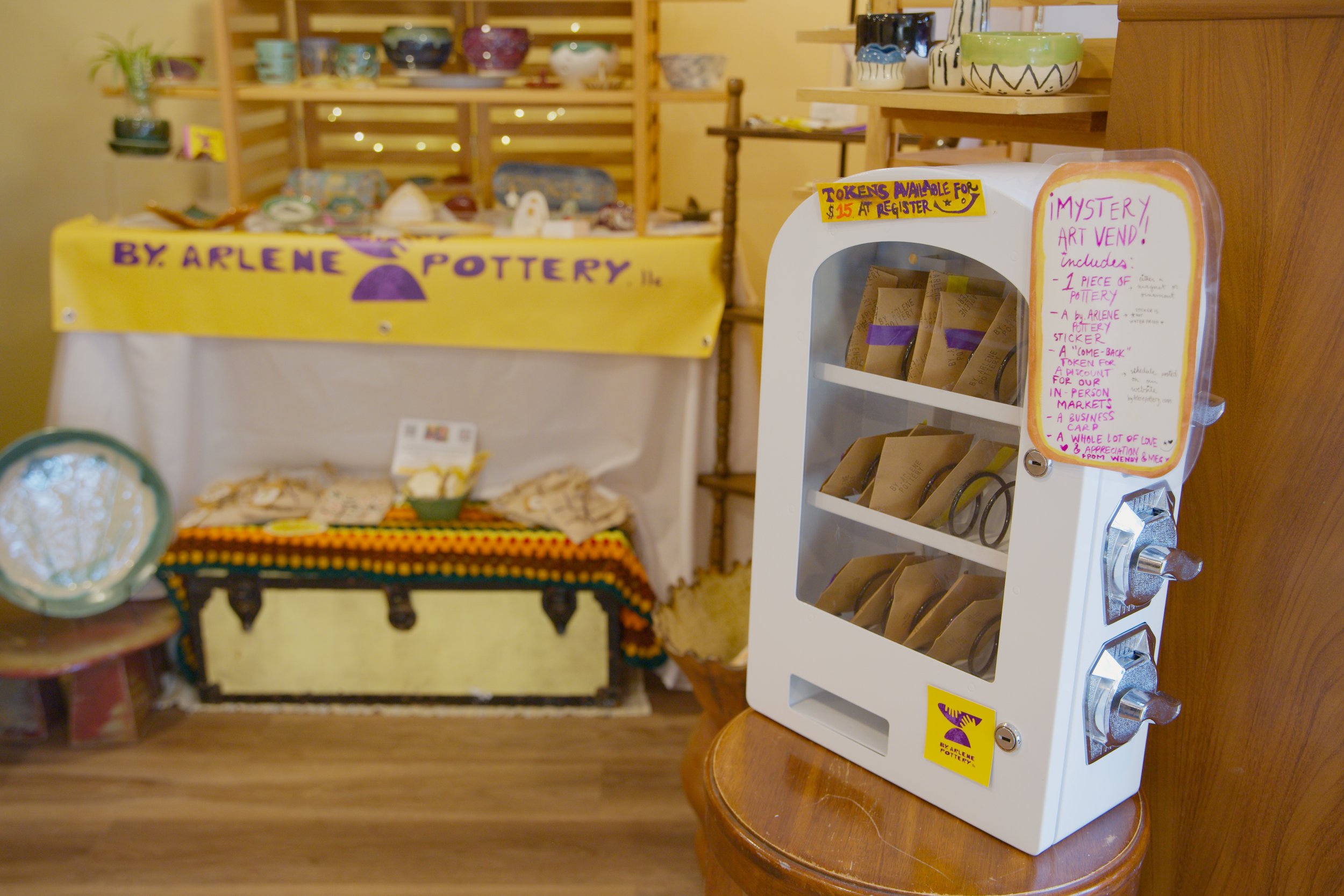 An Art vending machine with bags of pottery tokens, alongside a display table with pottery items, in a craft shop.