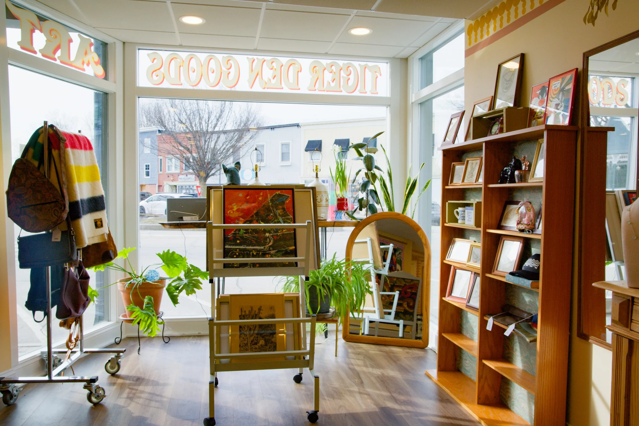 Inside a secondhand store with large front windows, displaying books, framed pictures, and plants on shelves and display stands, and a clothing rack with bags and scarves near the window.