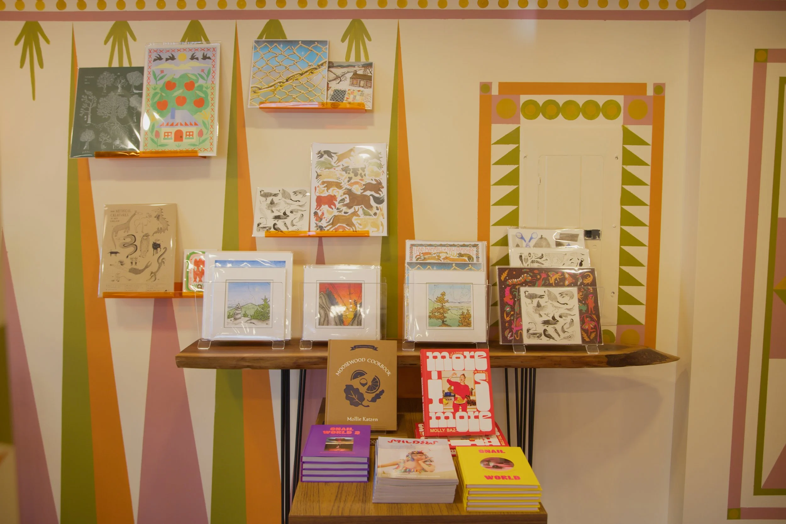 Display of various books and stationery on shelves against a painted wall with geometric and nature-themed patterns.