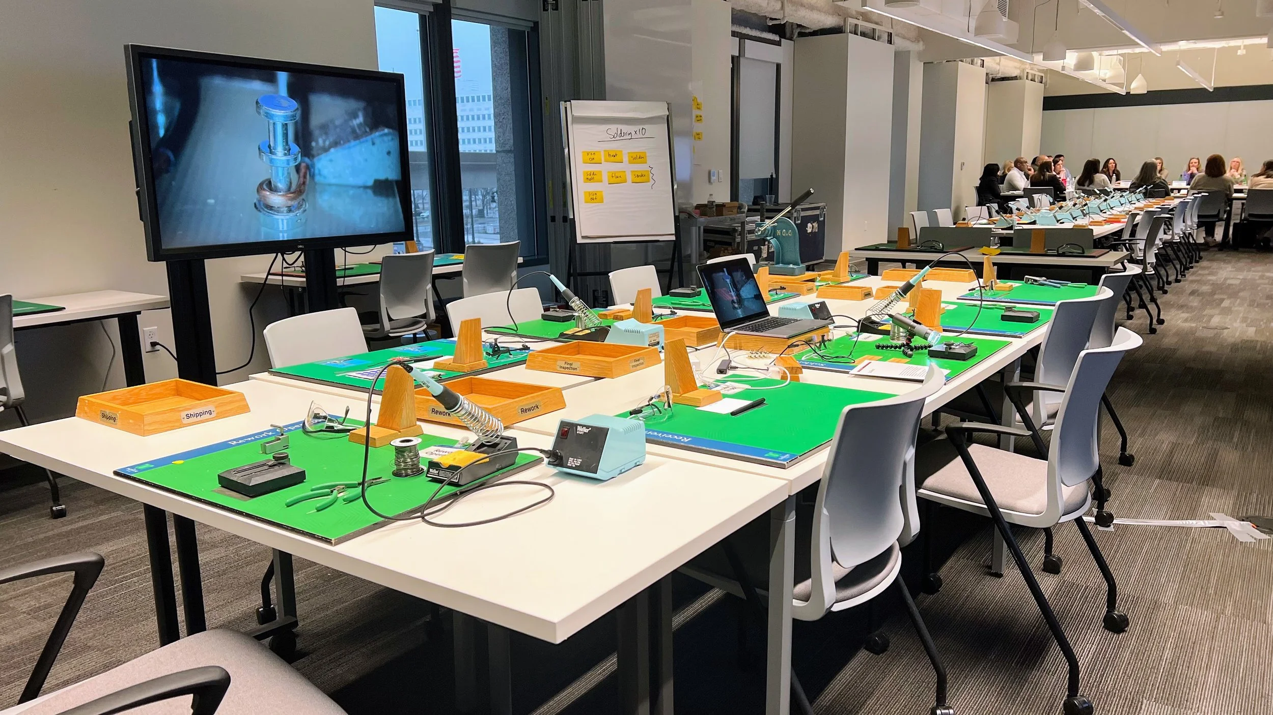 Conference room set up for a soldering event with workstations, soldering irons, tools, and large screens, with a group of people seated at a long table in the background.