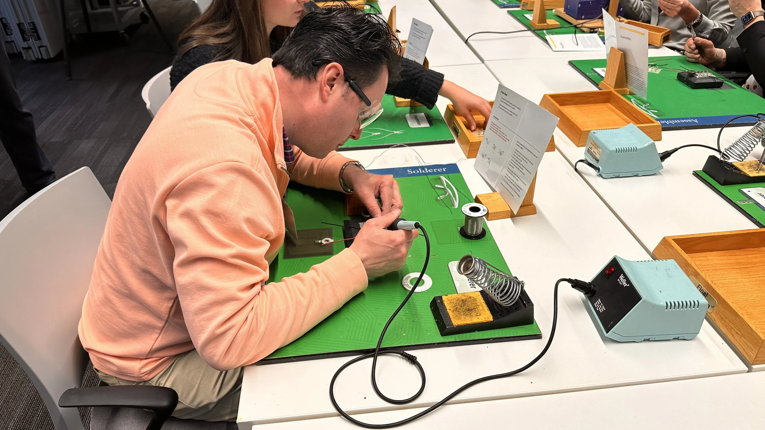 A man soldering electronic components at a workshop table with others, using a soldering iron and equipment, with several soldering stations, instructional signs, and tools on the table.