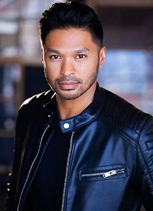Theatrical Acting headshot, wearing a black leather jacket, standing indoors with a blurred background.