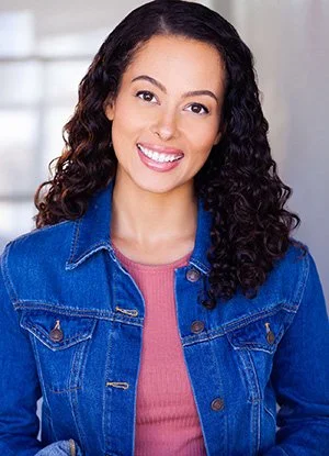 Young woman with curly hair smiling, wearing a denim jacket and pink shirt indoors.
