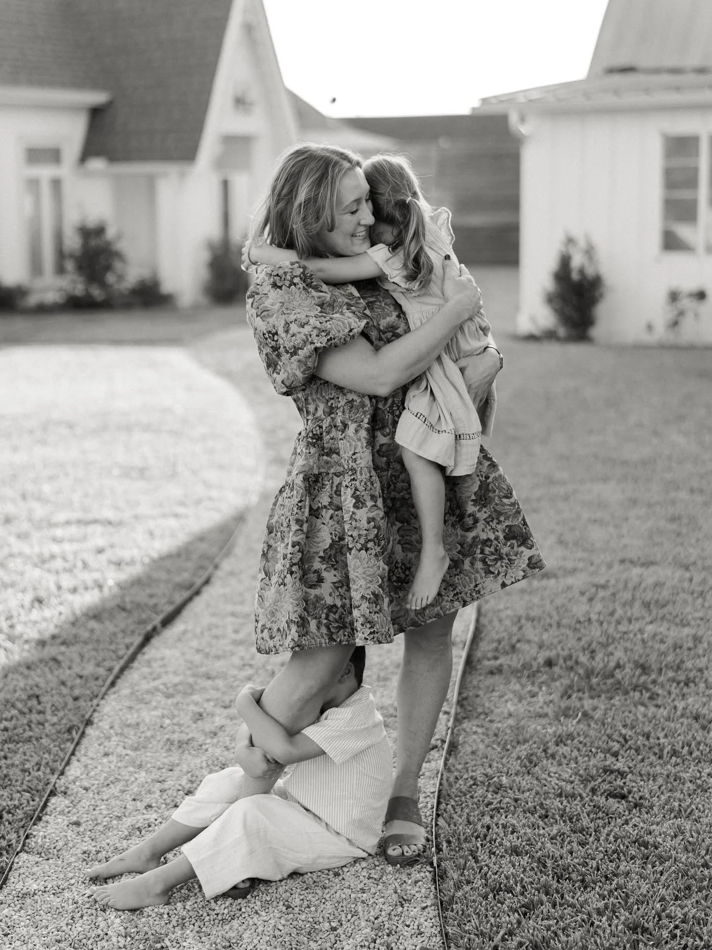A woman hugging two young children, one child is climbing on her while the other is sitting on the ground hugging her leg, on a grassy area with houses in the background.